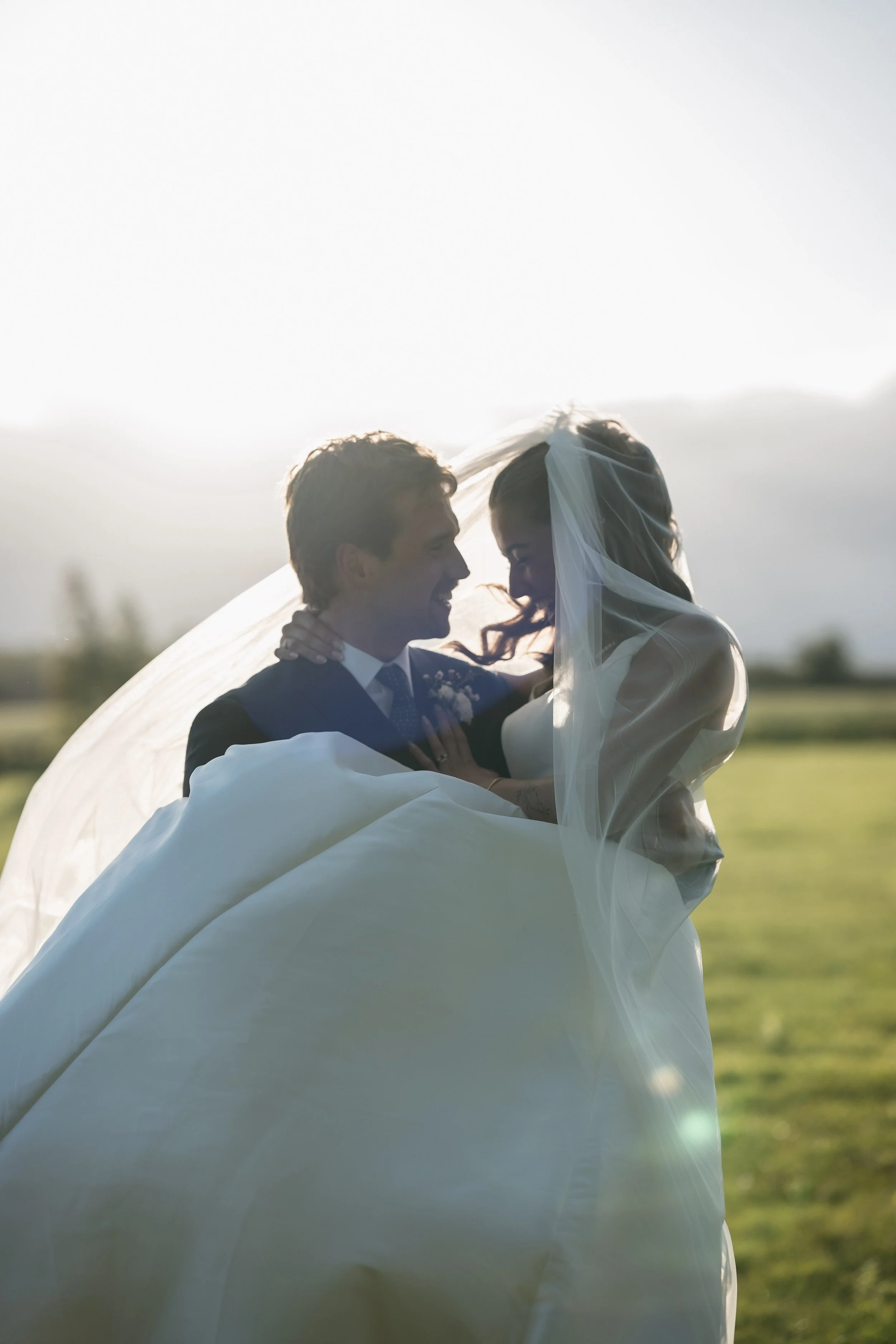 A bride and groom on their wedding day, standing outdoors with sunlight behind them, sharing a joyful moment under the bride's veil.