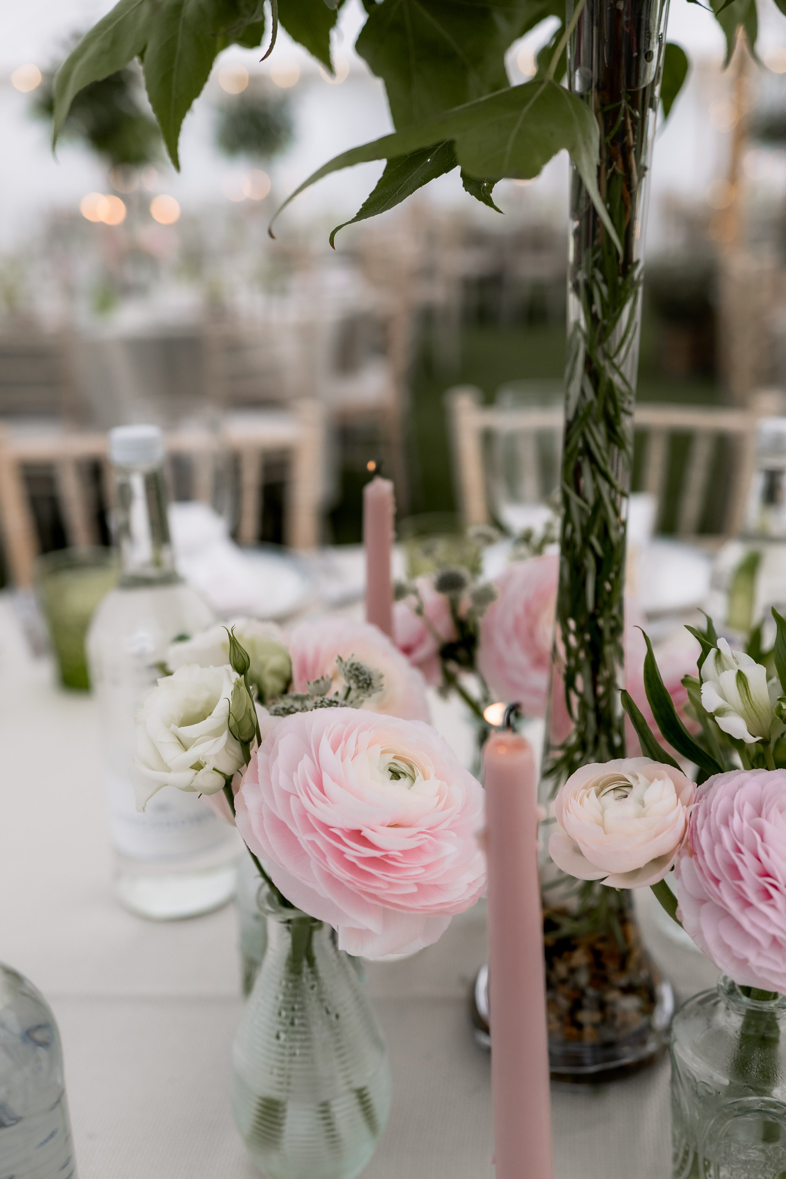 A close-up of a table centerpiece with pink and white flowers, glass vases, pink candles, and a blurred outdoor background.