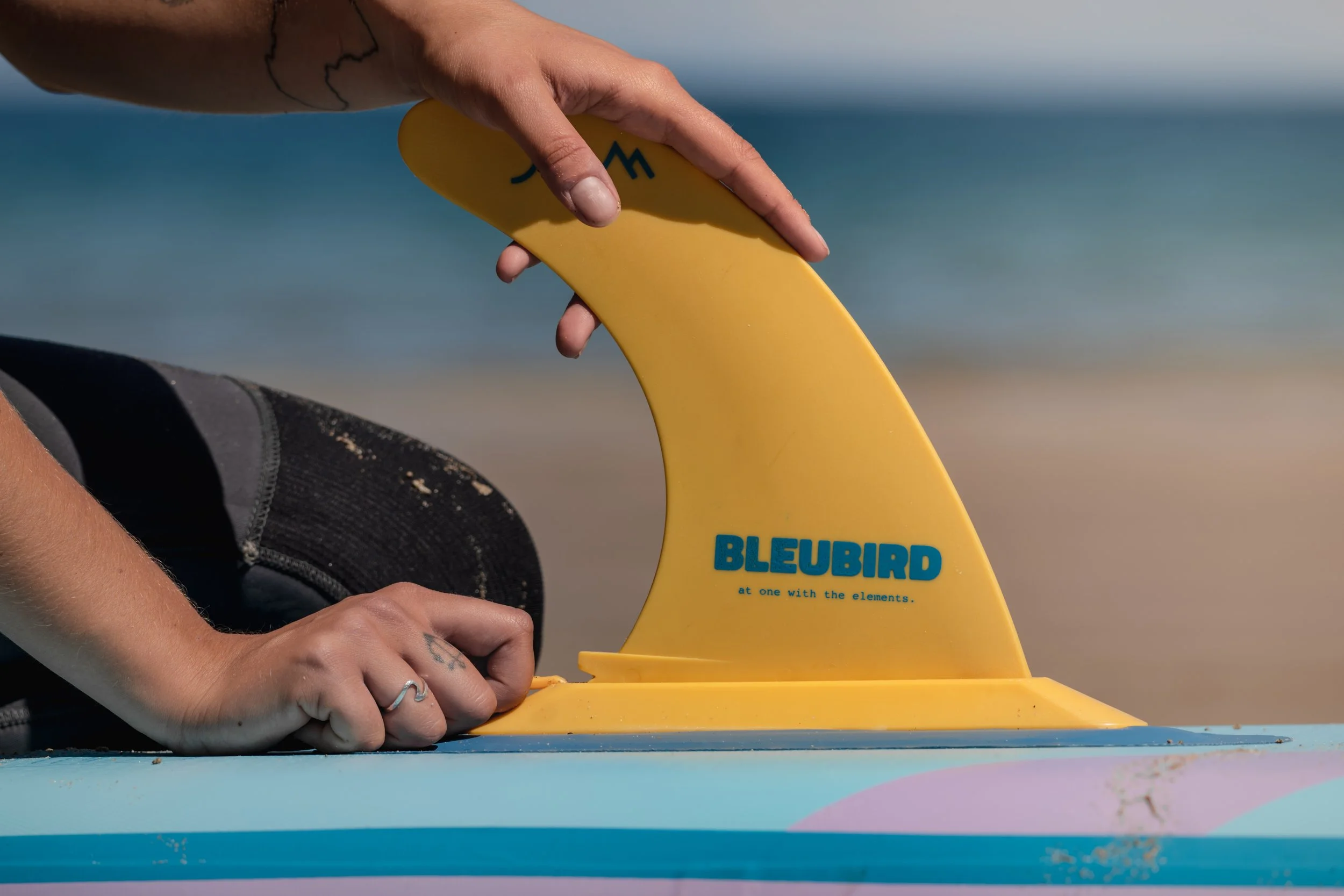 Close-up of a person fixing a yellow surfboard fin labeled 'BLEUBIRD' on a colorful surfboard at the beach, with sand and ocean in the background.