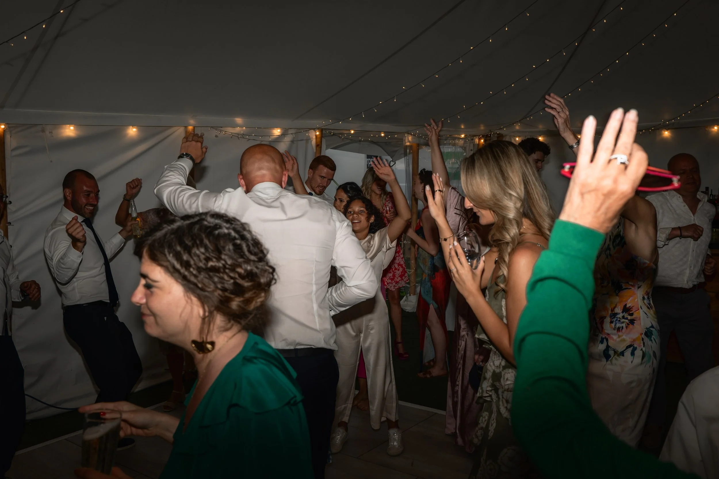 People dancing and celebrating at a party under a tent with string lights.