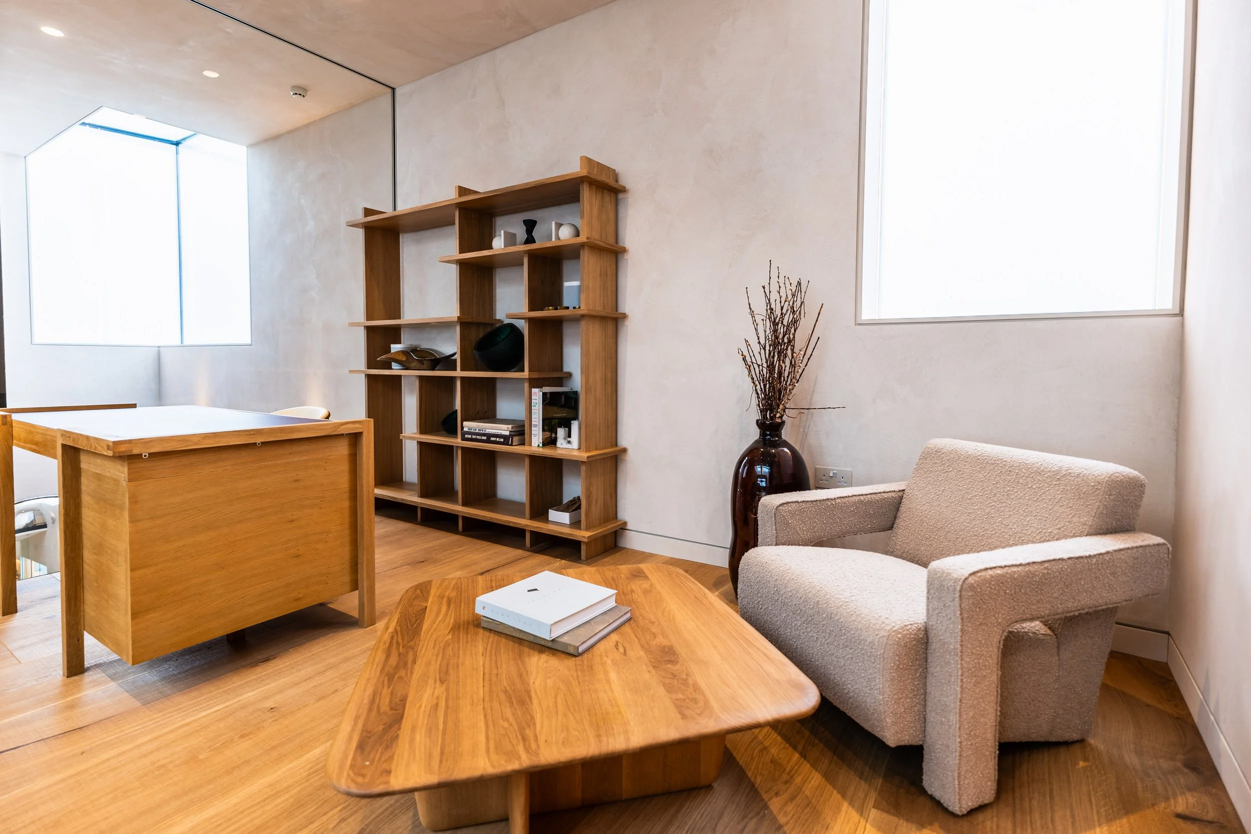 A cozy corner of a room with a beige armchair, a wooden coffee table with books, a tall wooden shelf with decorative objects, a large brown vase with dried branches, and two windows letting in natural light, all featuring minimalist design and neutra