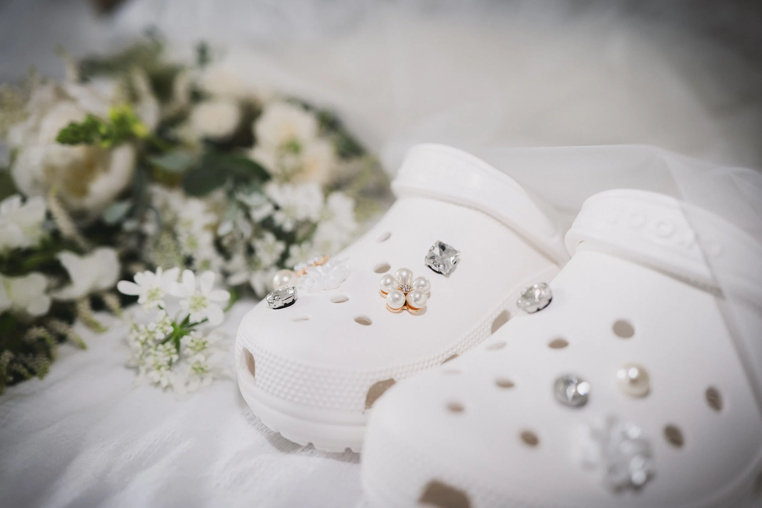 White children's slip-on shoes decorated with rhinestones and pearls, placed on a white cloth with small white flowers and a blurred bouquet in the background.
