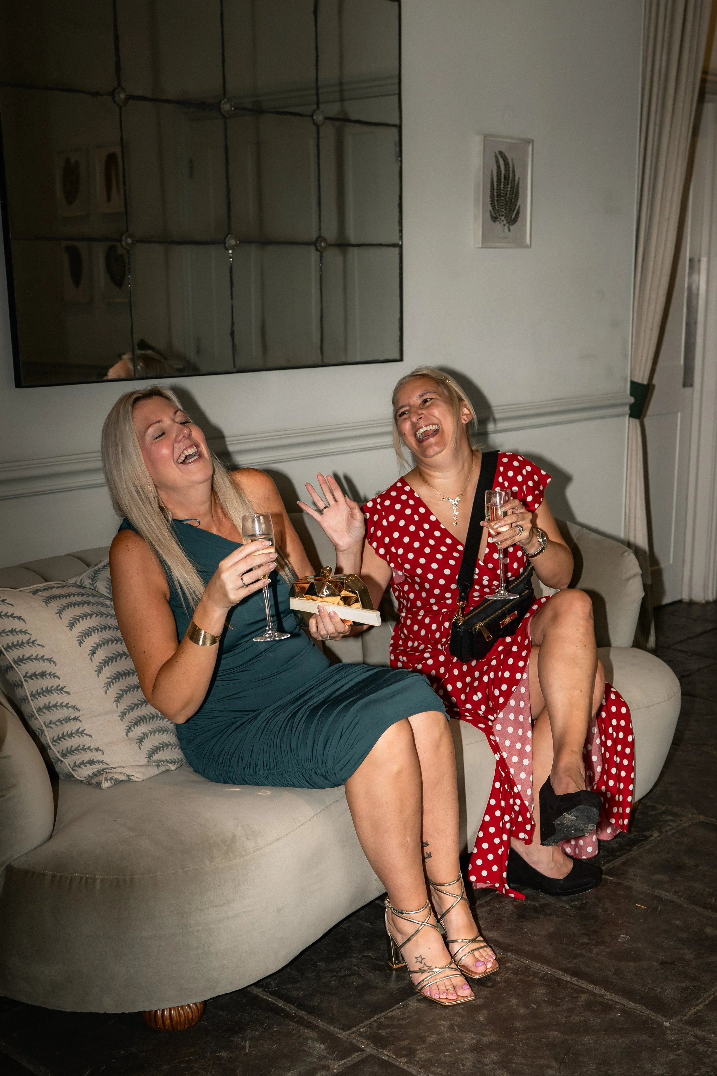 Two women sitting on a sofa, laughing and toasting glasses of champagne during a celebration.