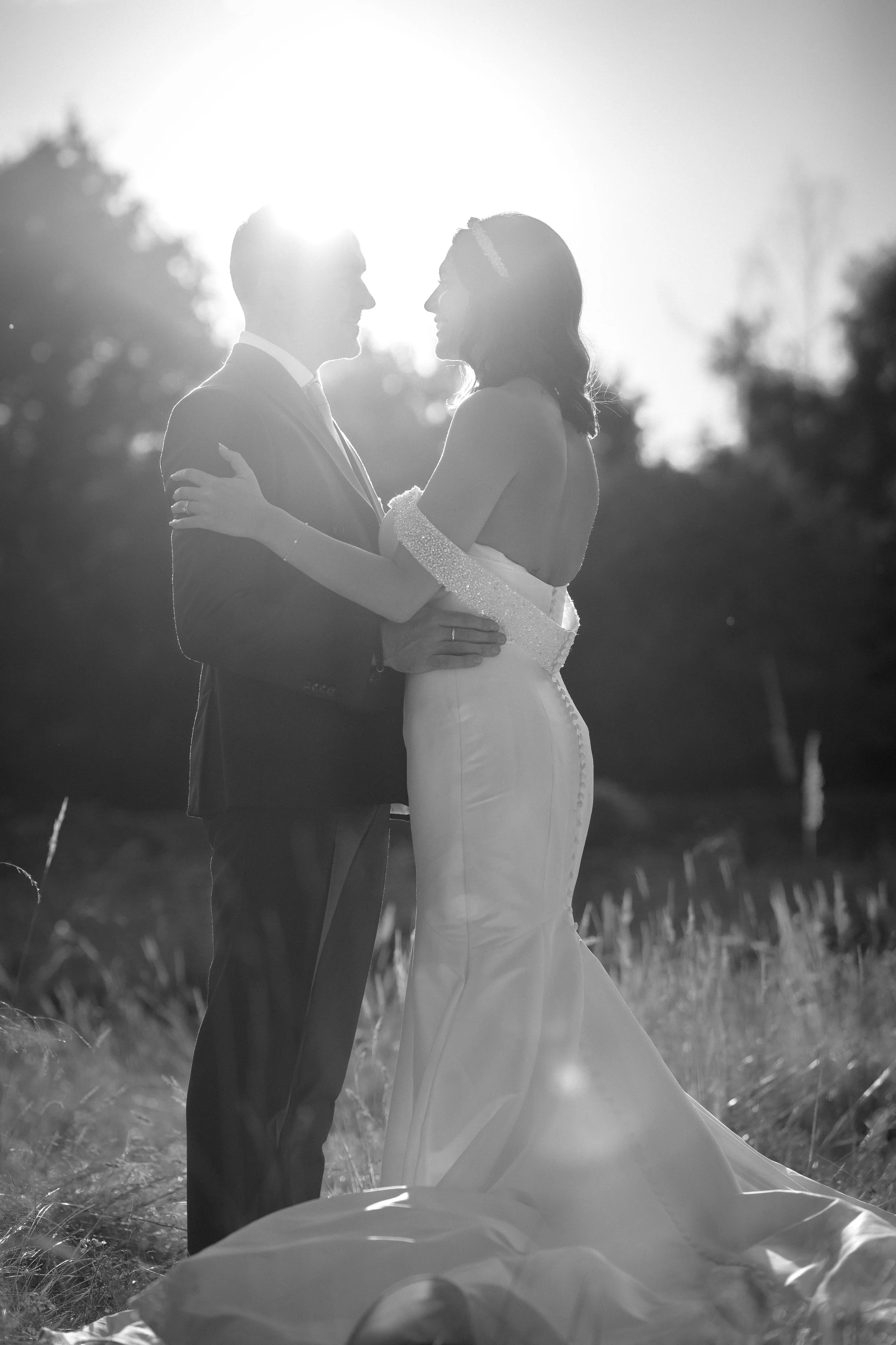 A black and white photo of a bride and groom standing outdoors, embracing and looking into each other's eyes, with the sun setting or rising behind them.