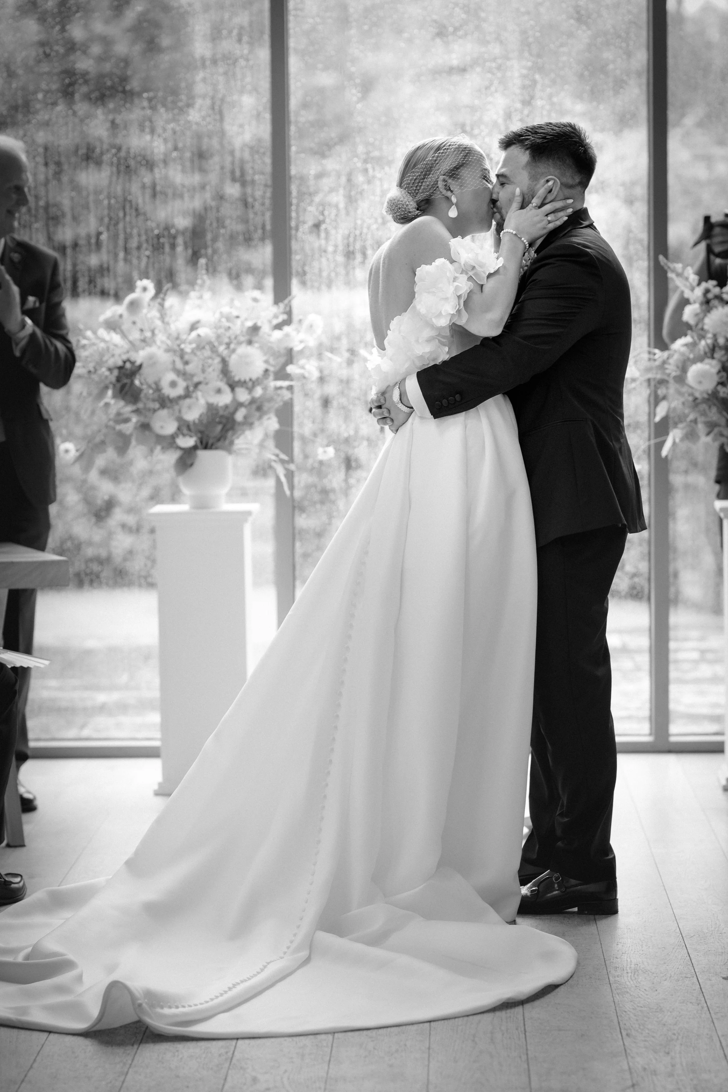 A bride and groom kiss during their wedding ceremony, with officiants and guests visible in the background, in front of large windows with rain outside.