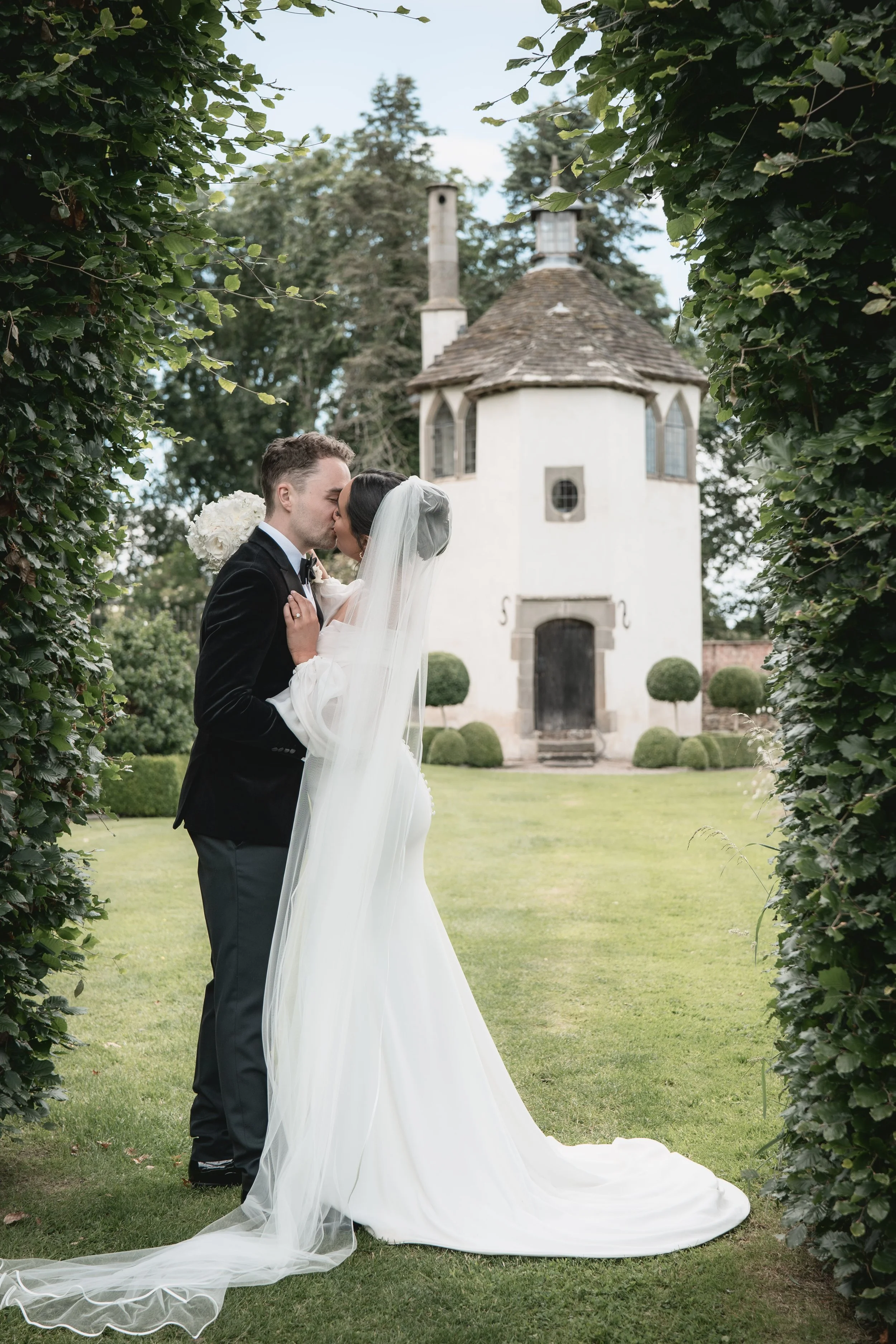 A bride and groom sharing a kiss outdoors on a wedding day, surrounded by greenery with a small white building in the background.