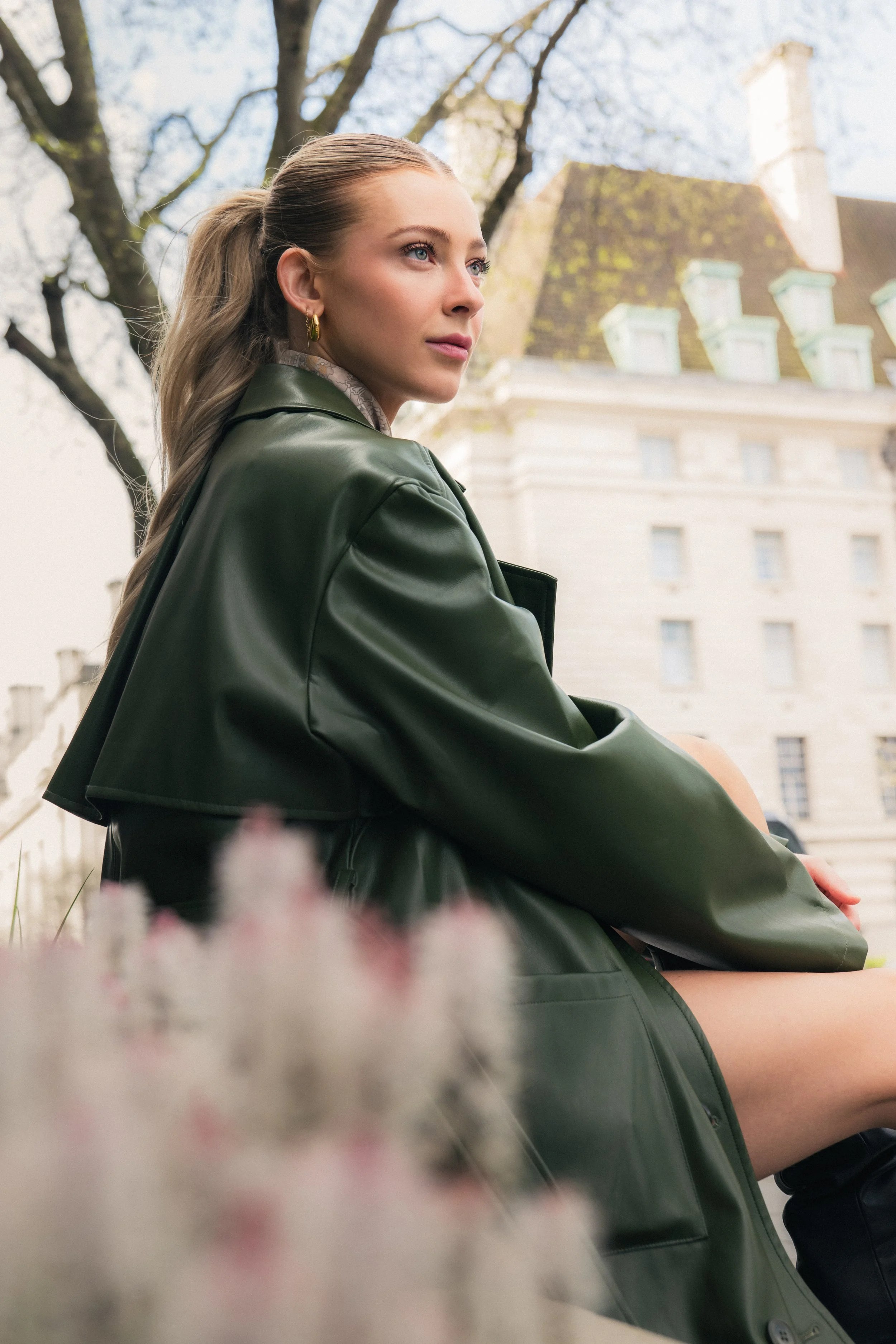 Young woman with blonde hair tied in a ponytail, wearing a dark green leather jacket, sitting outdoors near a building with multiple chimneys, trees, and a cloudy sky.