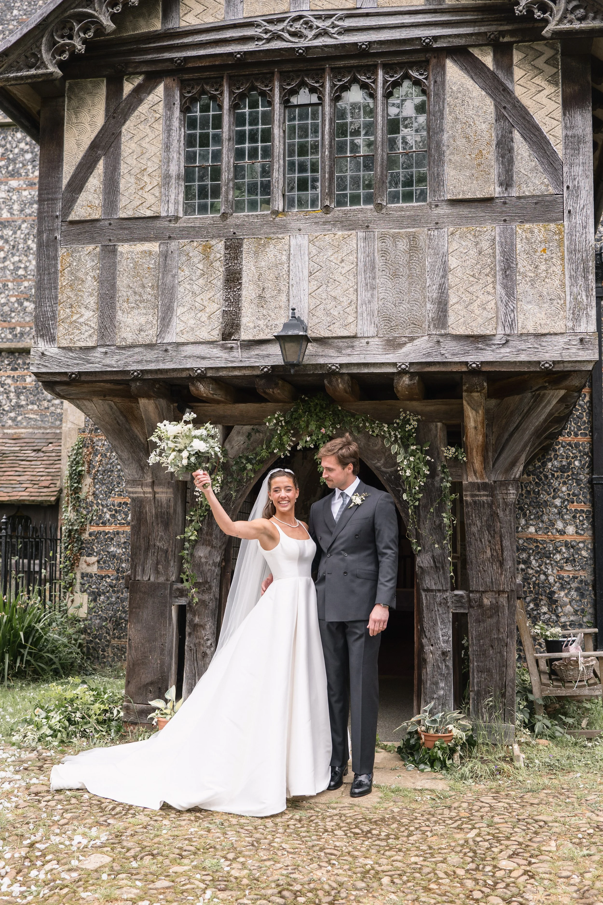 Bride and groom standing under a wooden arch at their wedding, with the bride holding a bouquet and smiling, and the groom looking at her.