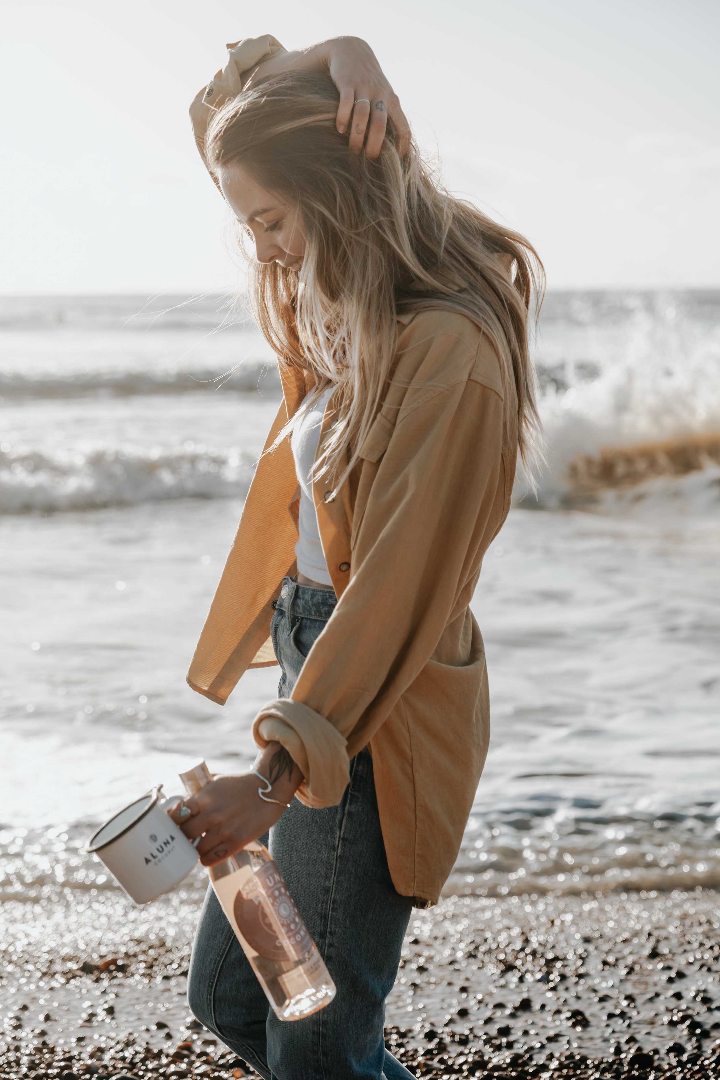 Woman standing on the beach near the ocean, holding a white mug and a clear water bottle, wearing a beige jacket, white shirt, and jeans, with her hair blowing in the wind.