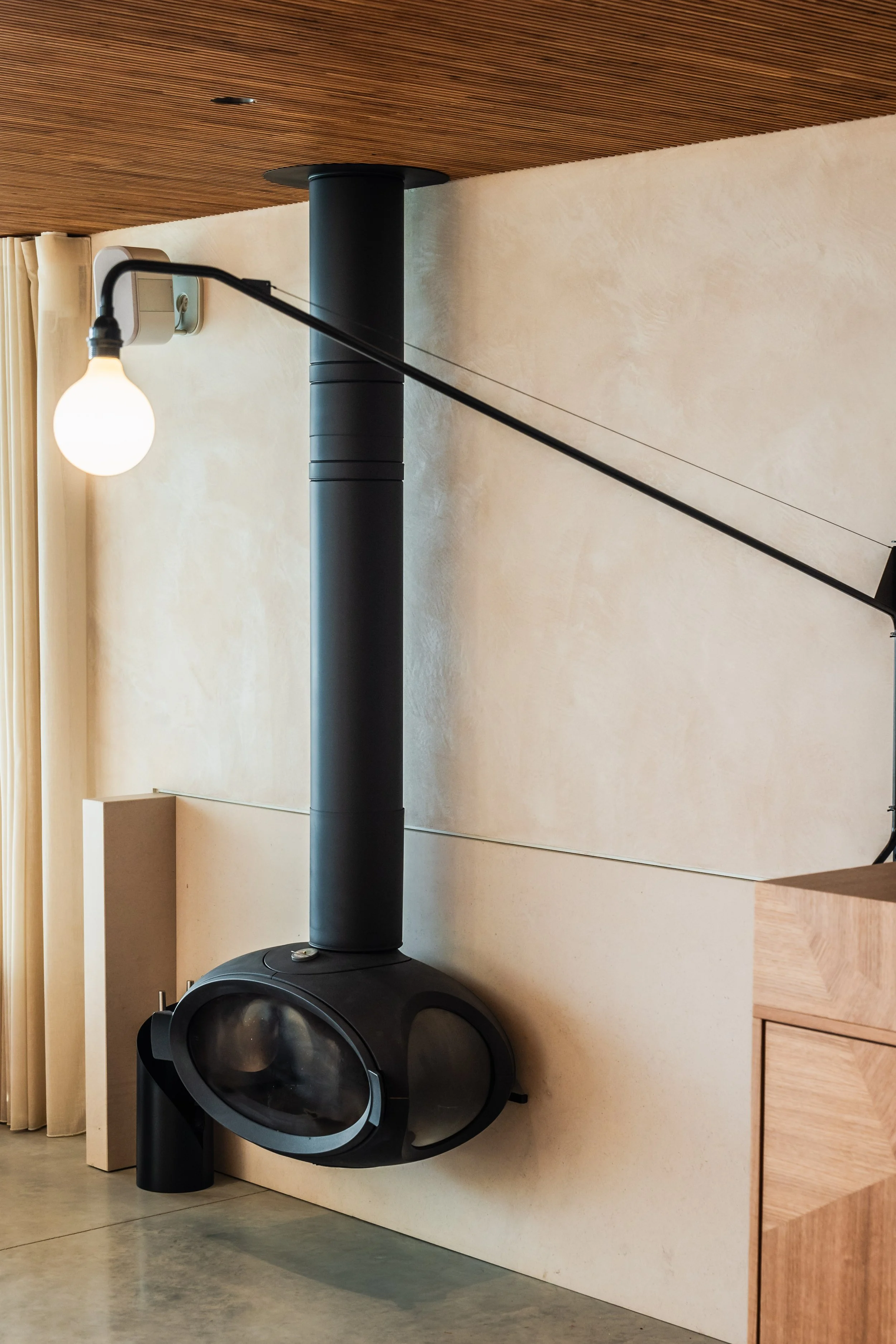 Modern black wood-burning fireplace with a tall chimney pipe, embedded in a beige wall, with a hanging light bulb and beige curtains in the background.