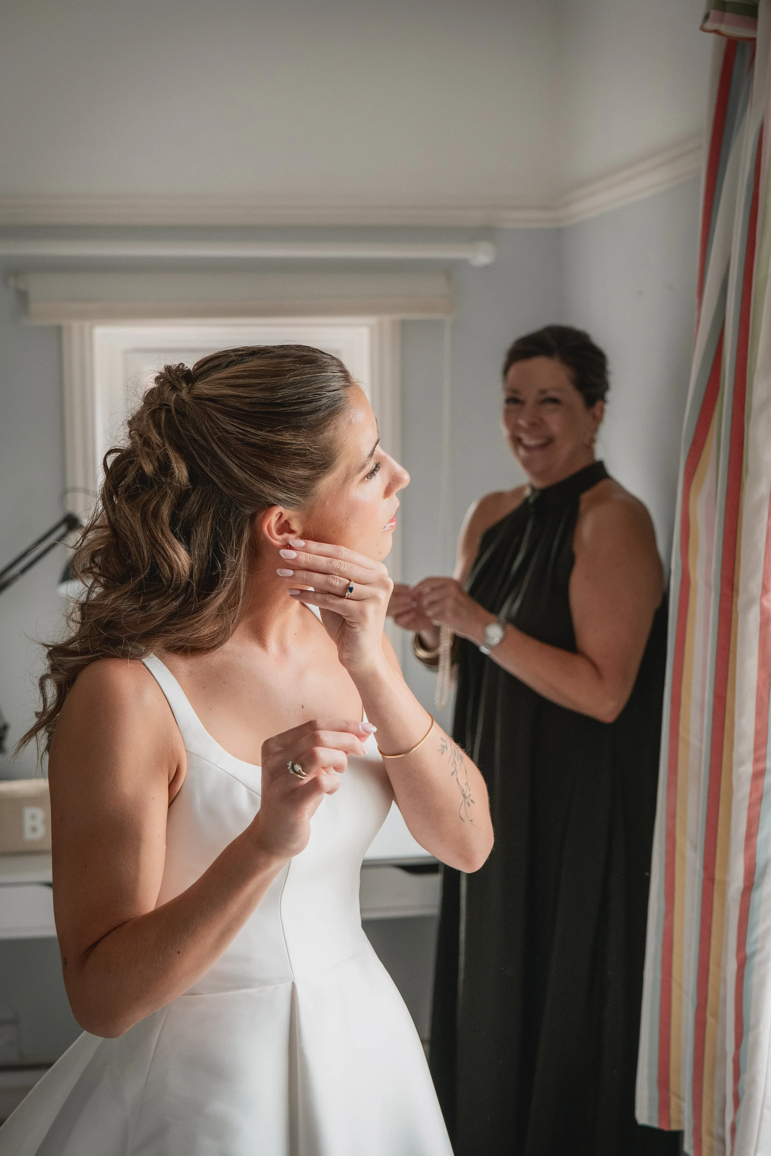 A bride in a white wedding dress prepares for her wedding as her mother helps with the final touches, standing in a softly lit room near a window with striped curtains.
