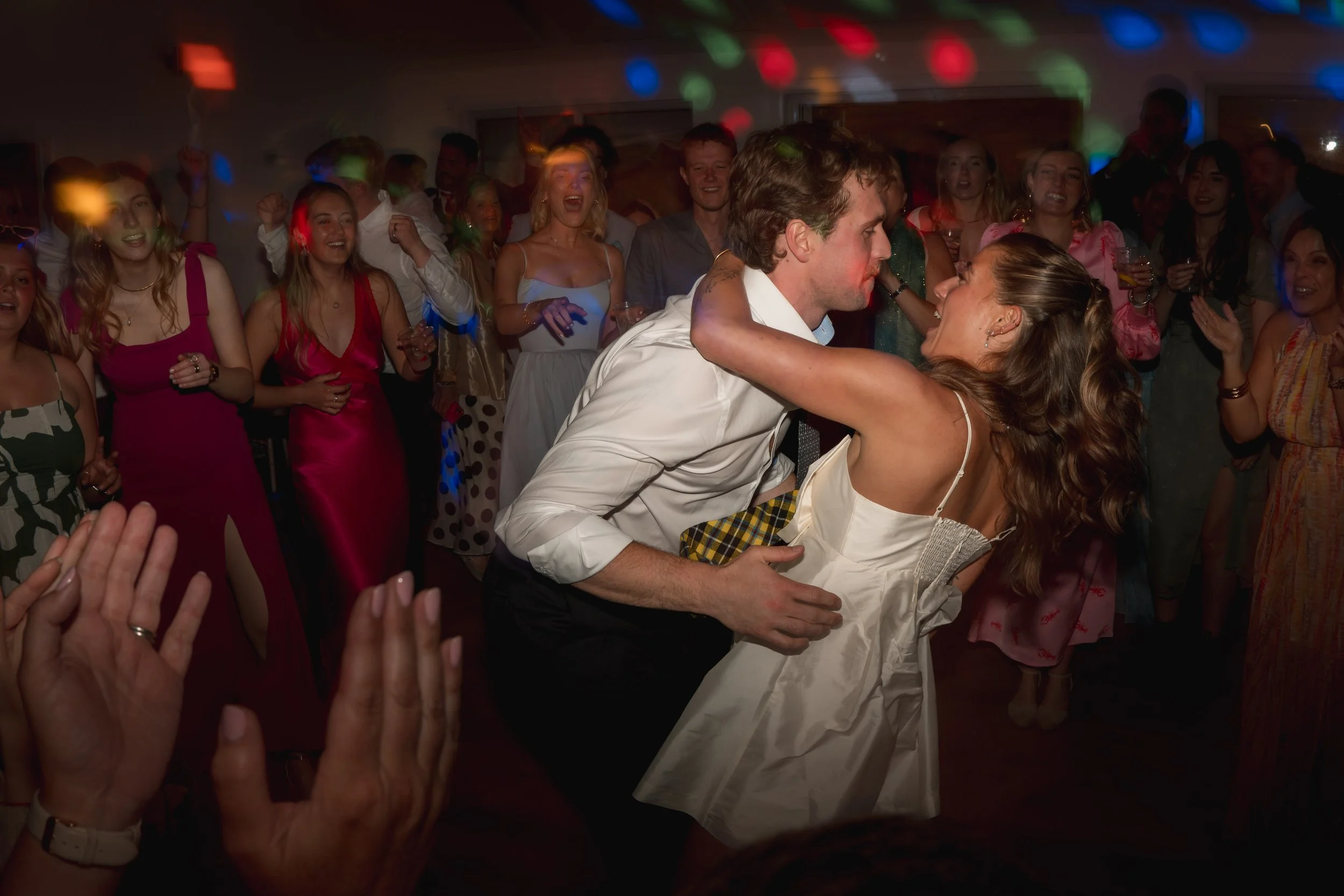 A couple dancing closely at a party surrounded by smiling and cheering friends in celebration.
