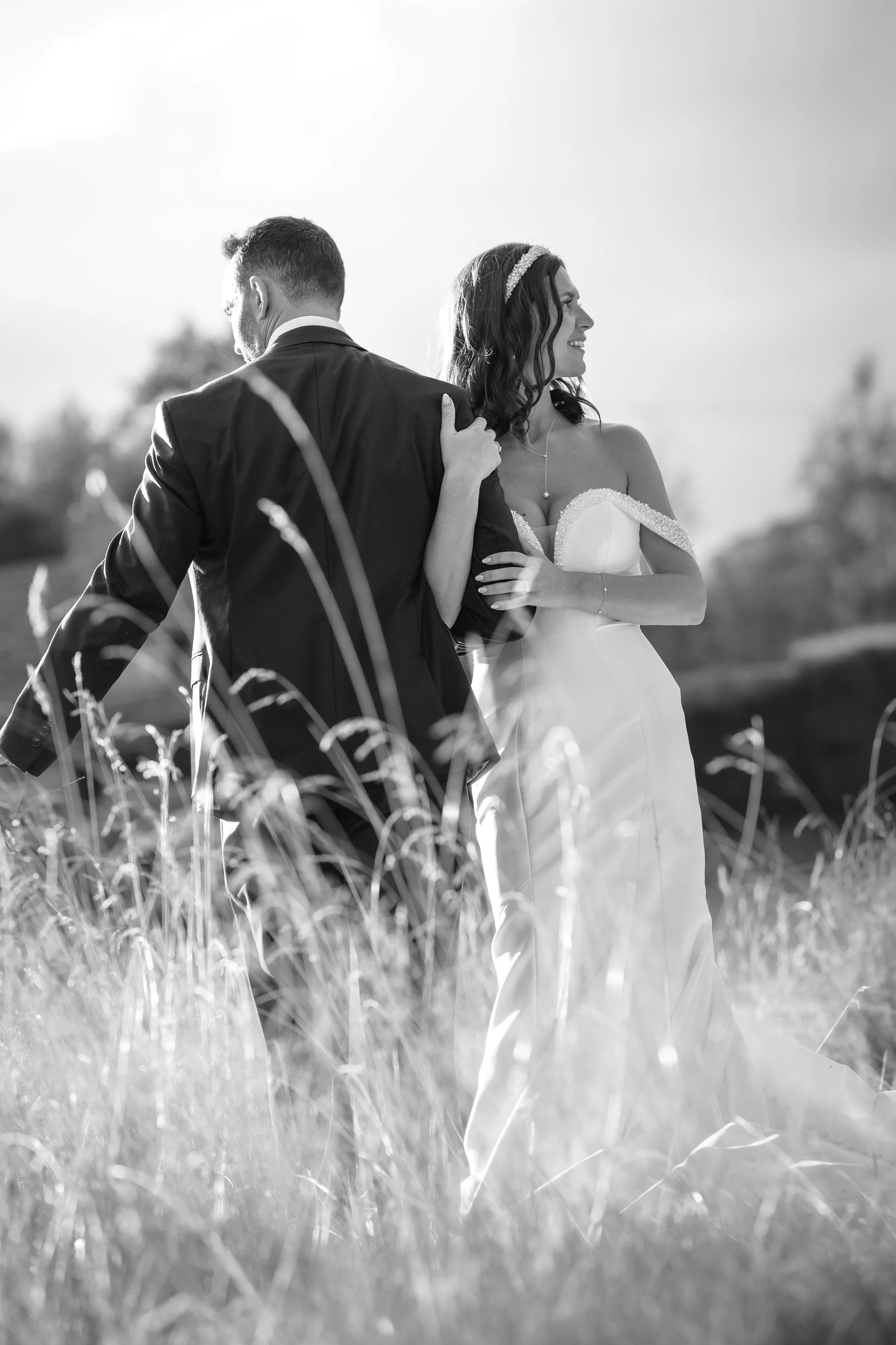 Black and white photo of a bride and groom standing outdoors in a field, embracing each other. The bride wears a strapless wedding gown with off-the-shoulder sleeves, and the groom is in a suit. They are smiling and looking away from each other.