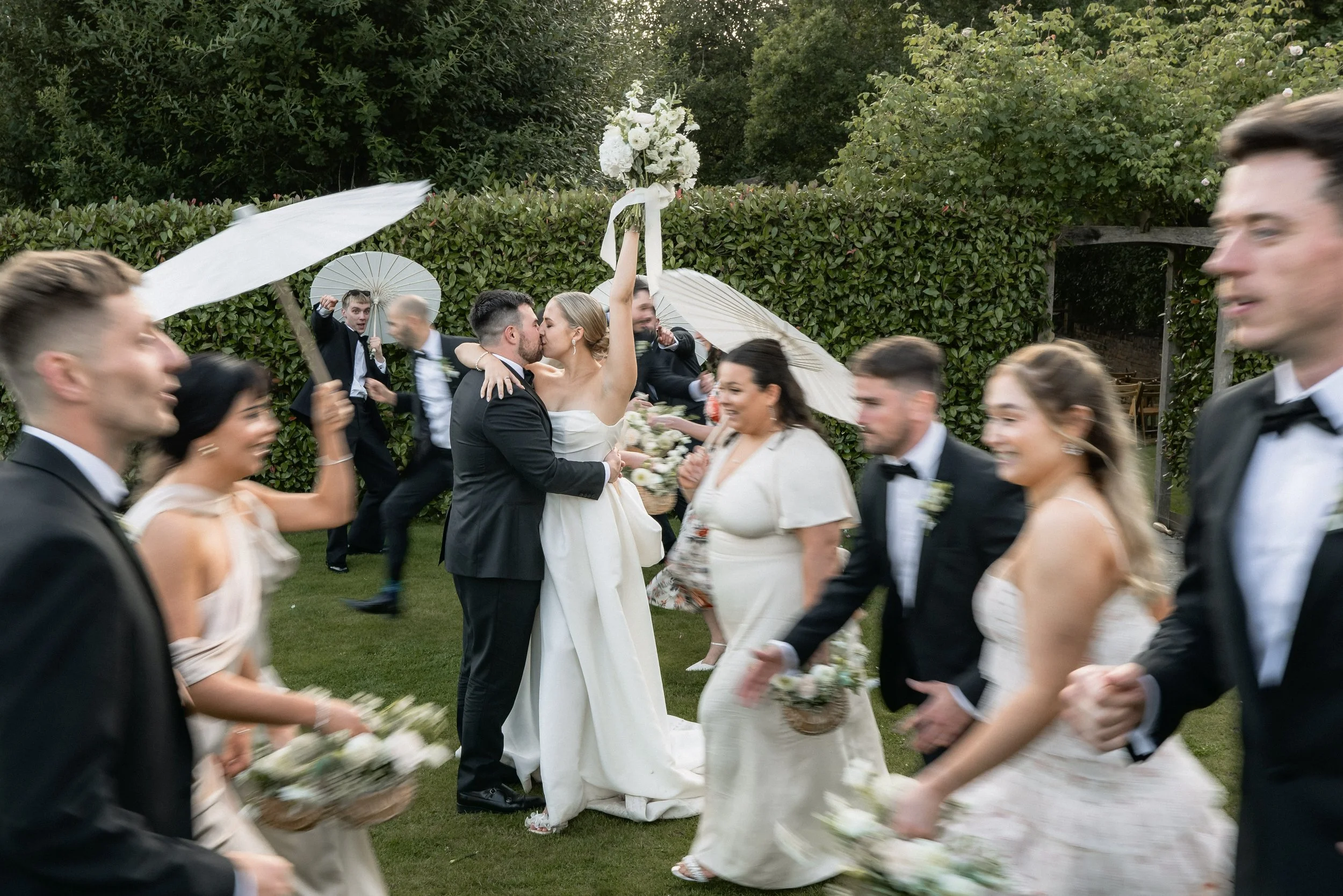 Wedding celebration with bride and groom kissing, surrounded by cheerful guests in formal attire, some holding umbrellas and flowers, outdoors in a garden.