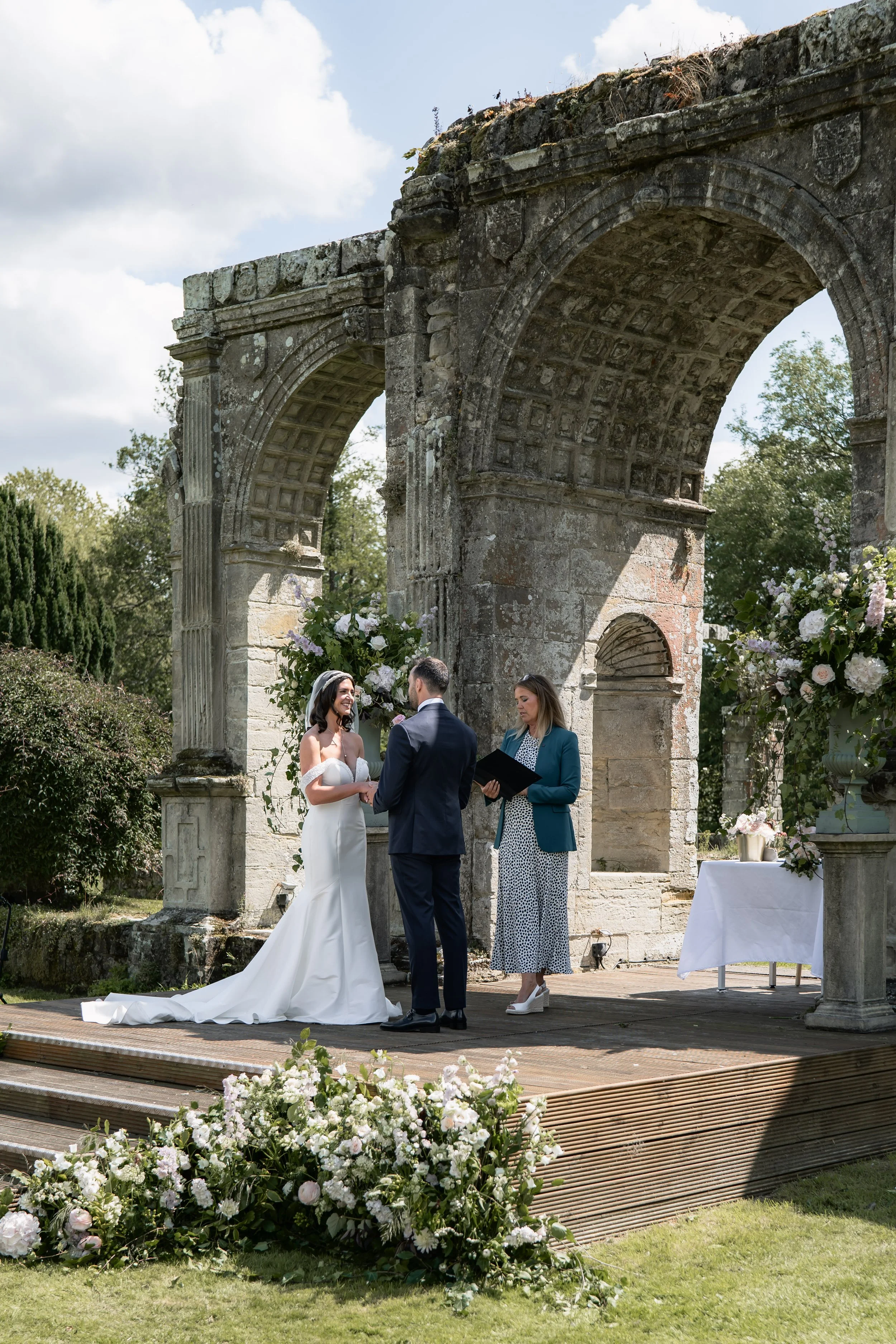 A couple getting married outdoors under an ancient stone arch with floral decorations, officiant reading vows, wedding dress, and groom in suit, surrounded by lush greenery and bright sky.