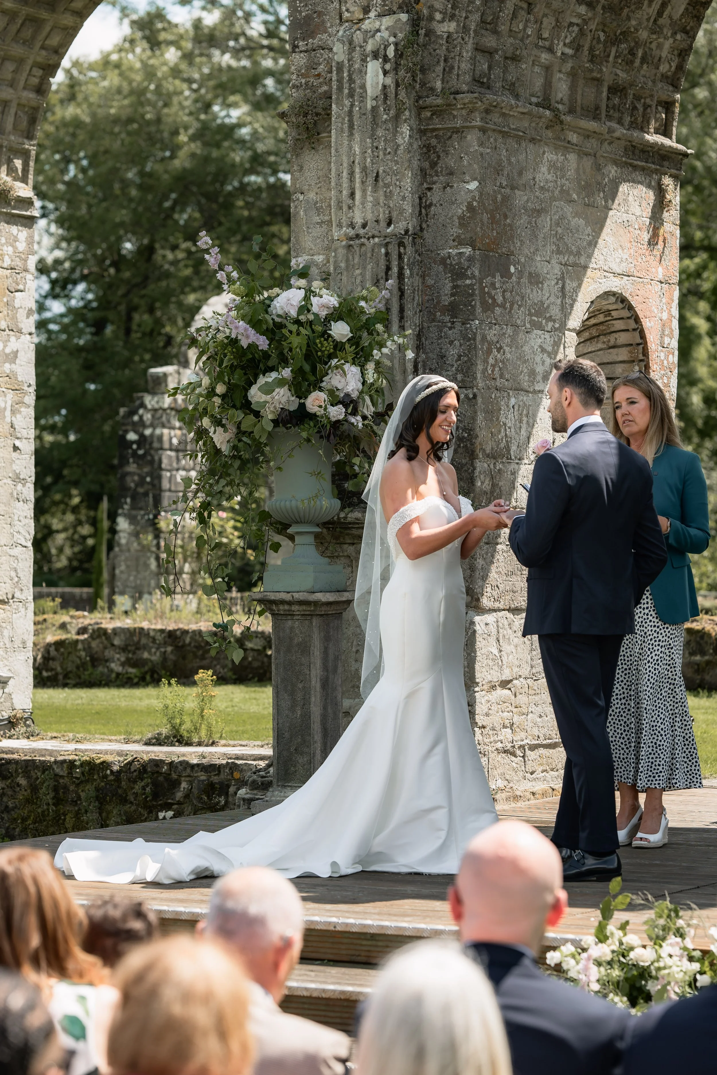 A bride and groom exchange vows during an outdoor wedding ceremony under a stone arch, with guests watching in the foreground and a large floral arrangement on the side.