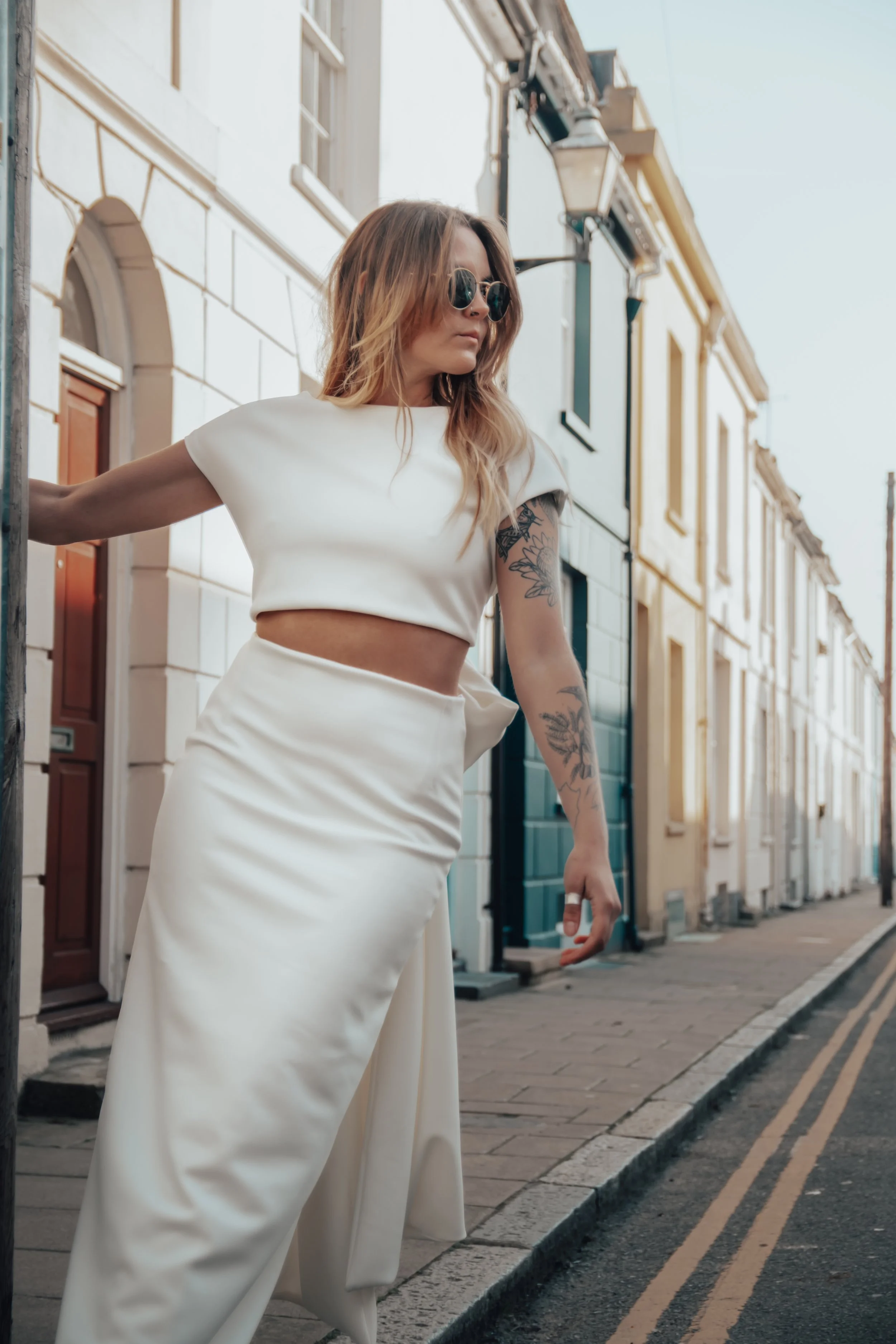Fashionable woman with tattoos wearing sunglasses, a white crop top, and high-waisted white pants, standing on a city sidewalk.