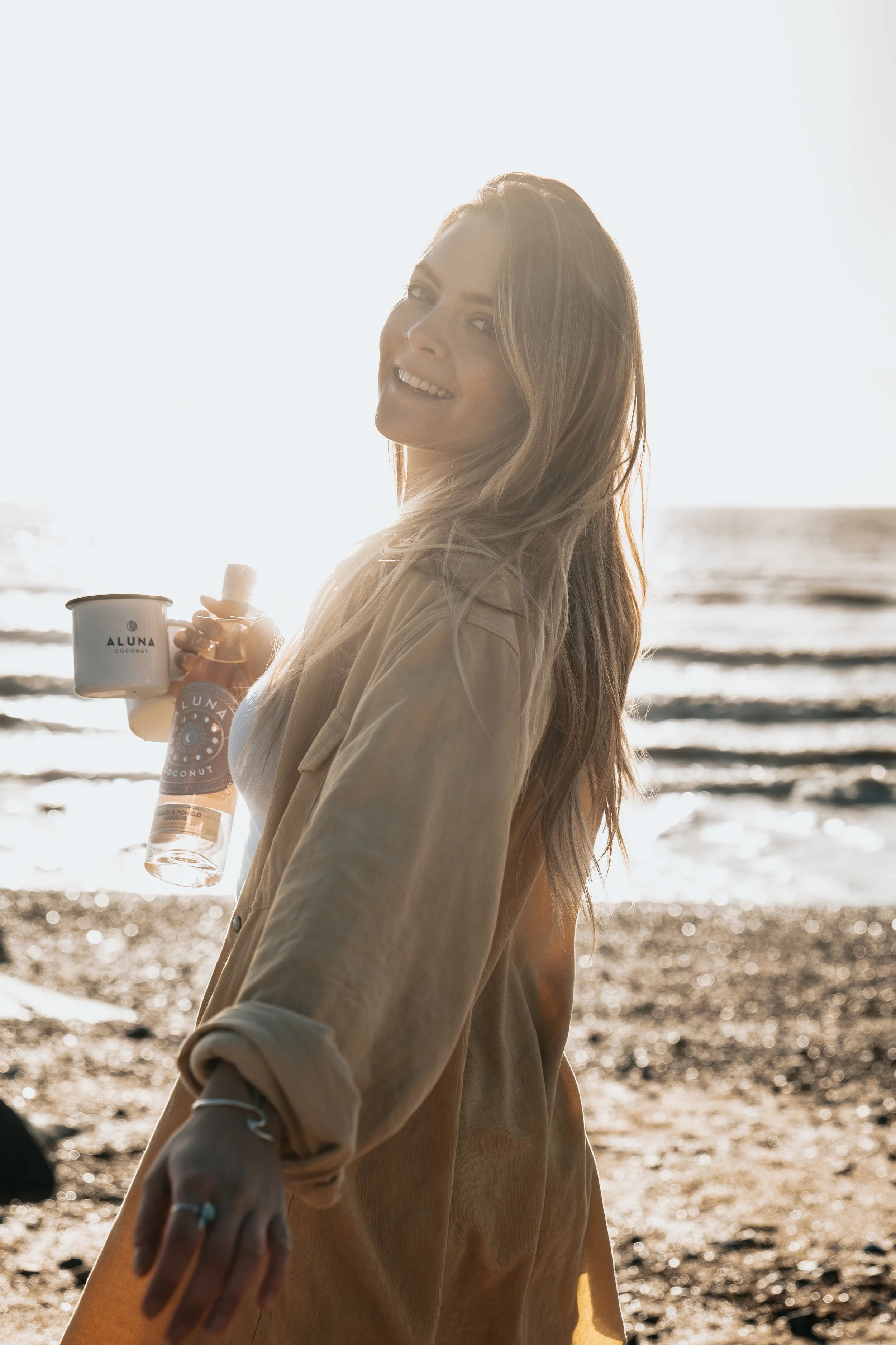 Woman smiling on the beach holding a coconut drink and a bottle, backlit by the sunset.