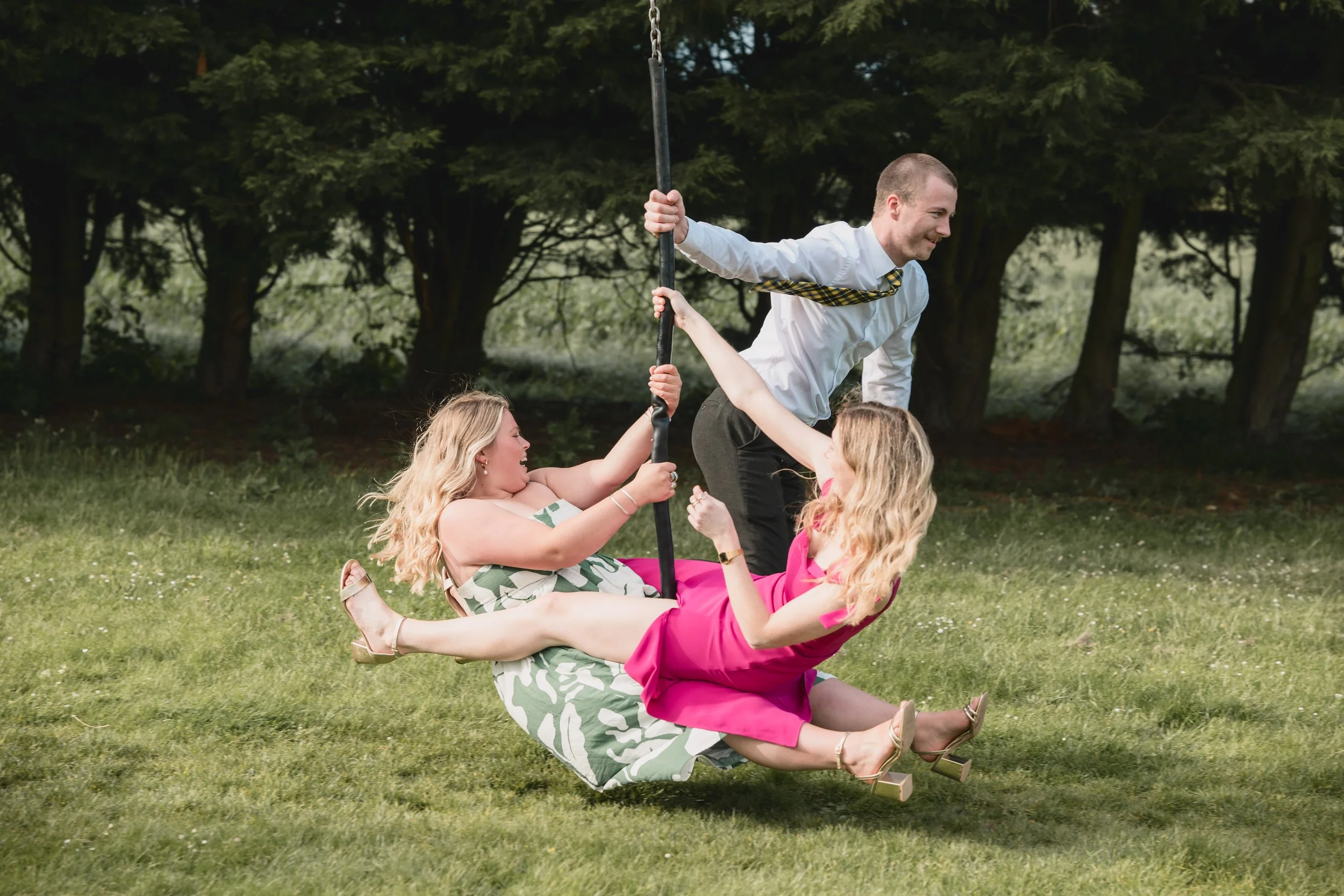 Two women in dresses on a swing set, one in a pink dress, the other in a patterned dress, playing with a man in formal attire, outdoors with trees in the background.