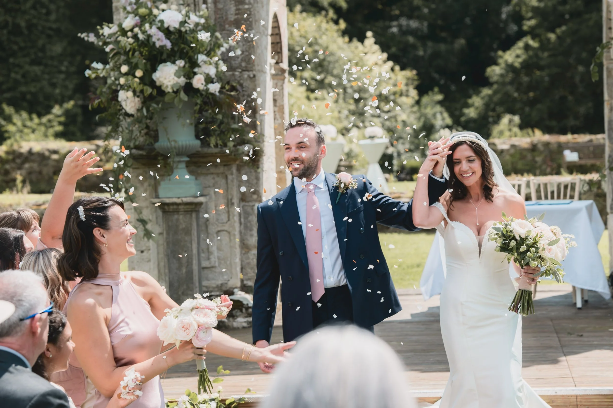 A newlywed couple celebrating at their outdoor wedding reception, with confetti being thrown, surrounded by friends and family, in a garden setting with greenery and stone structures.