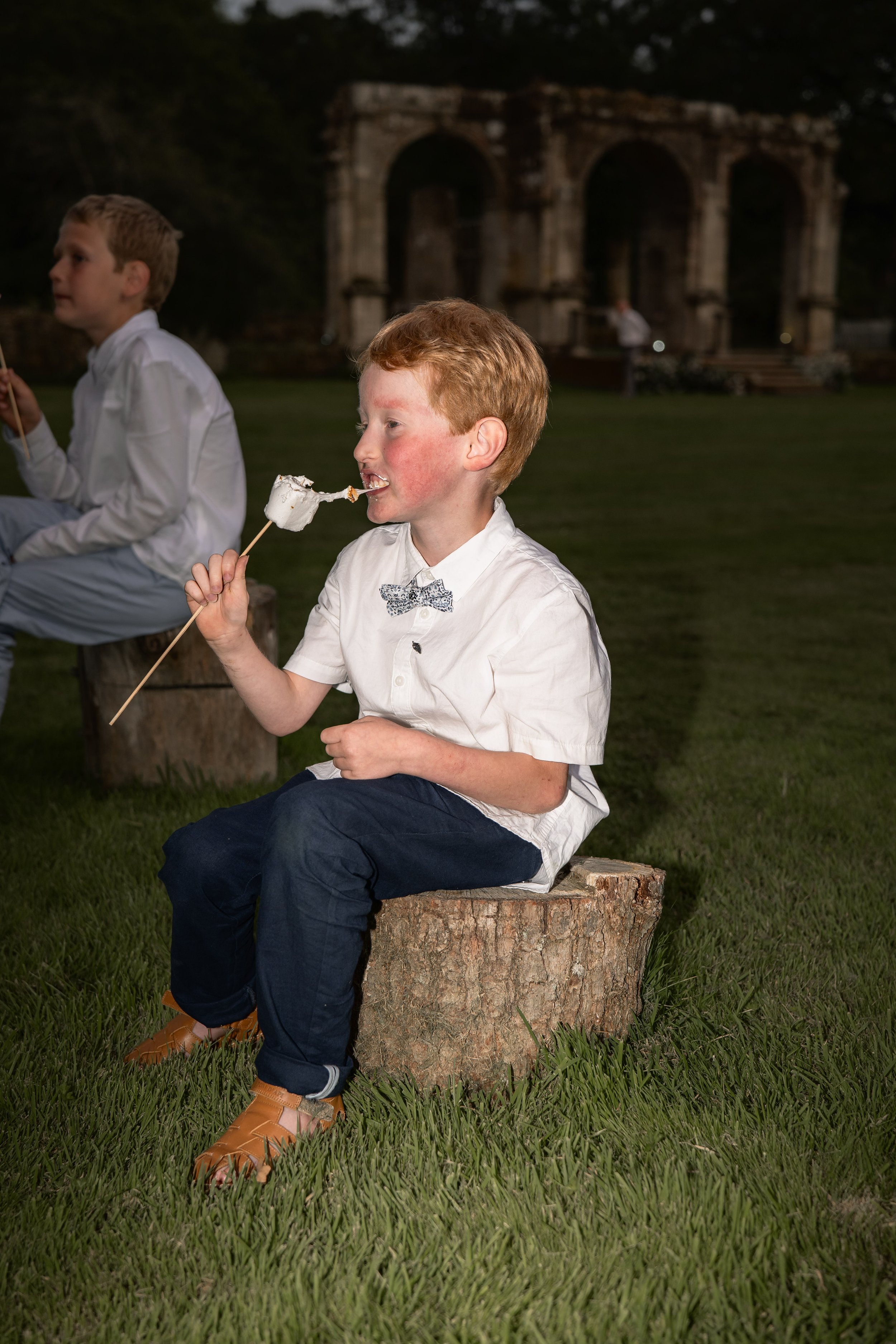 A young boy with red hair and freckles, dressed in a white shirt with a bowtie and dark pants, is sitting cross-legged on a tree stump at an outdoor event during the evening. He is eating a marshmallow on a stick, with another child in similar attire