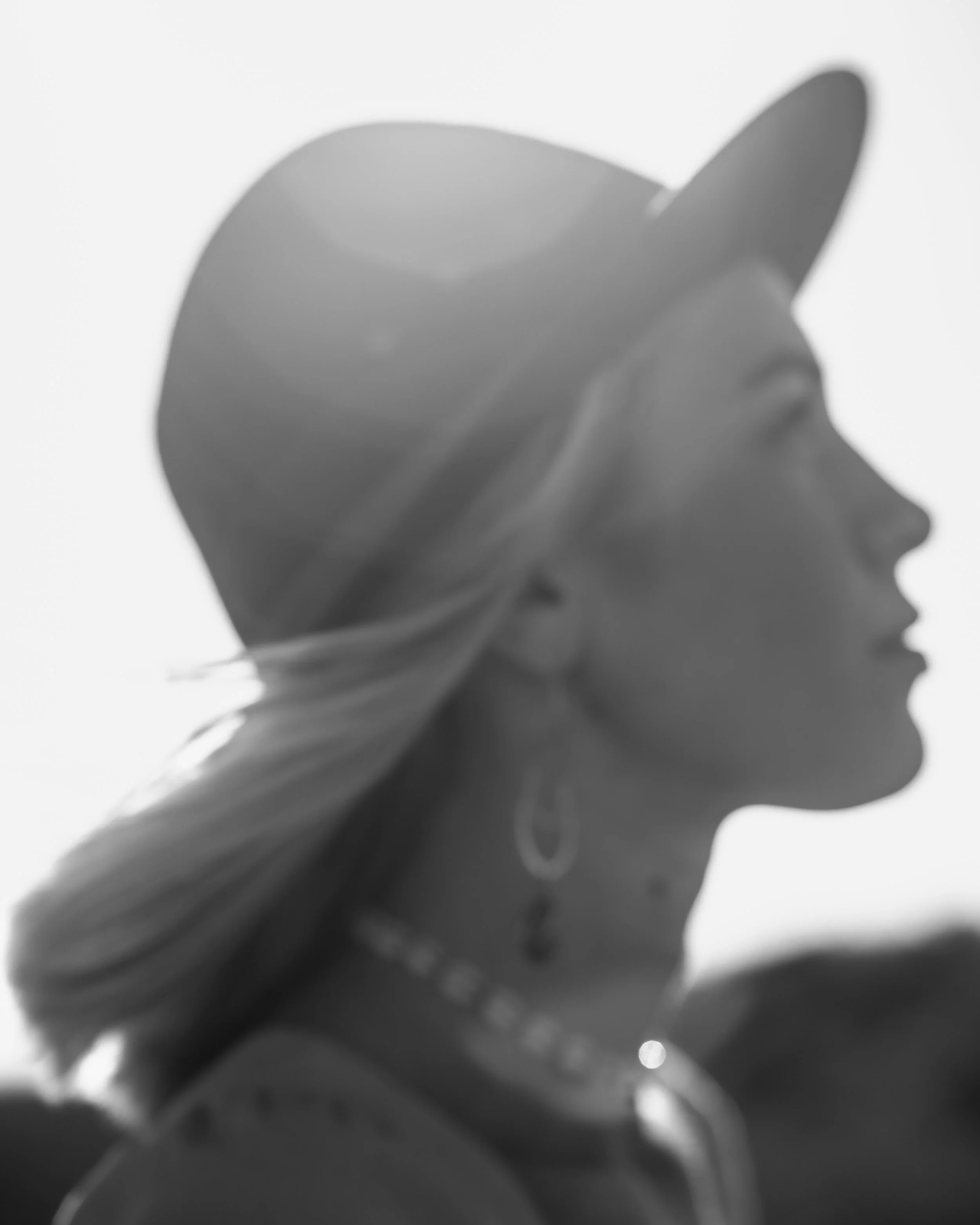 Side profile of a woman wearing a wide-brimmed hat and earrings, with her hair flowing in the wind, in black and white.