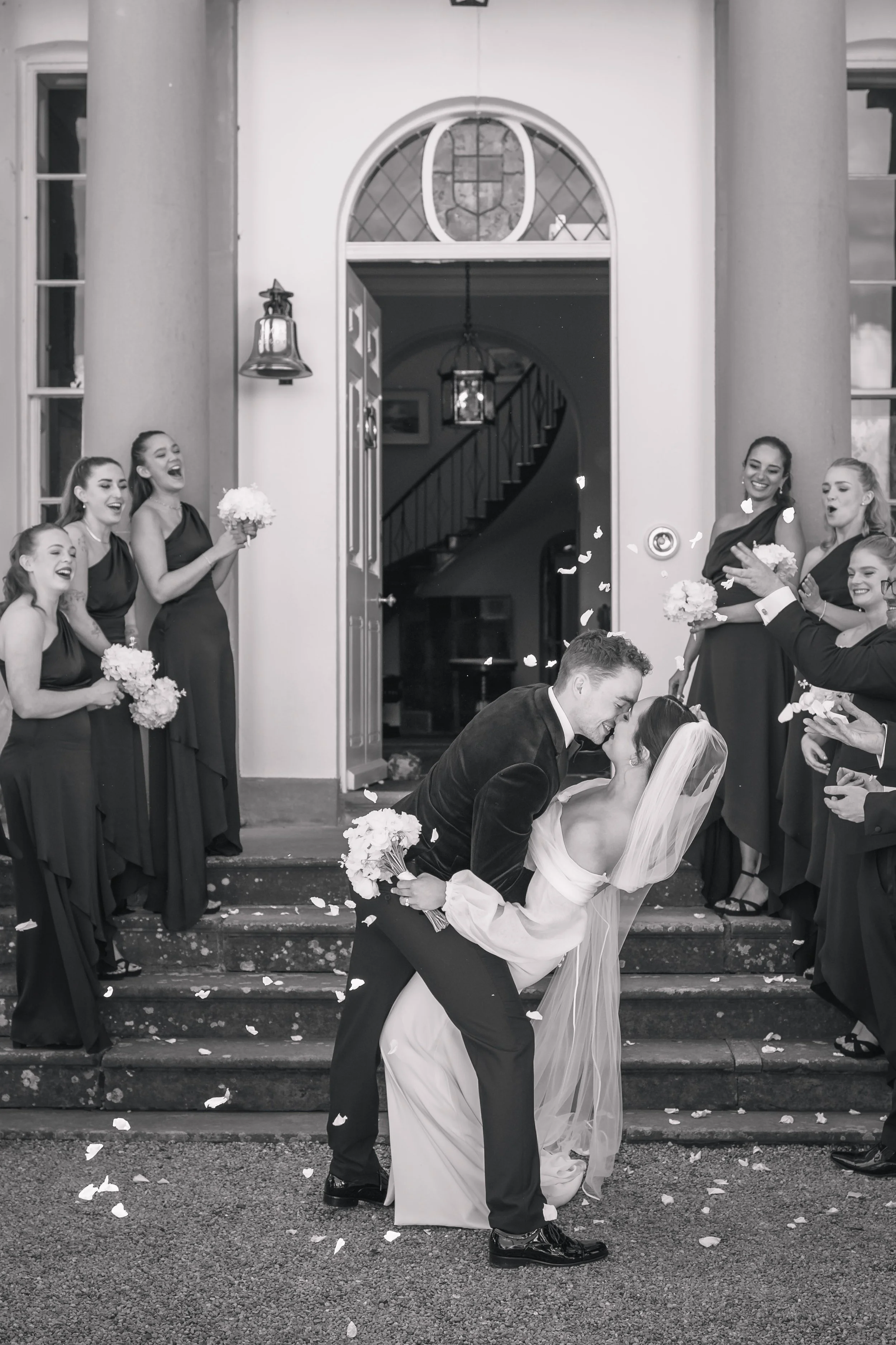 A black-and-white photograph of a newlywed couple sharing a kiss on the steps of a building, surrounded by friends in formal attire, with flower petals in the air, celebrating their wedding.