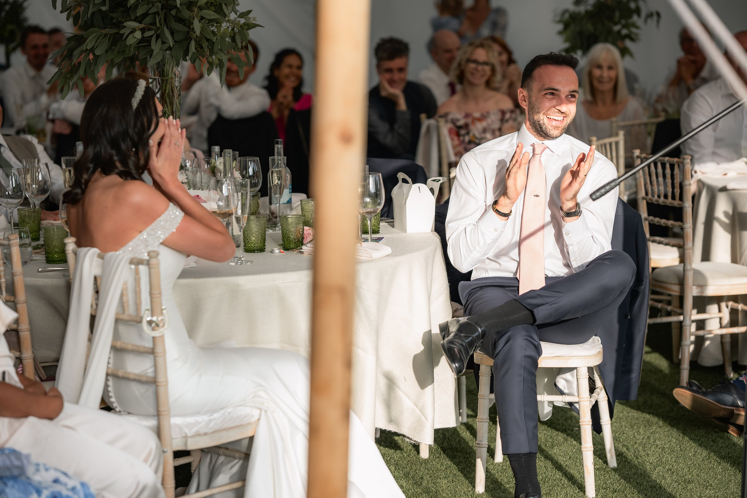 A man in a white shirt and pink tie is sitting and clapping, smiling at a wedding reception. A woman in a white dress with off-the-shoulder sleeves is sitting across from him, covering her face with her hands. Guests are seated at tables with glasses