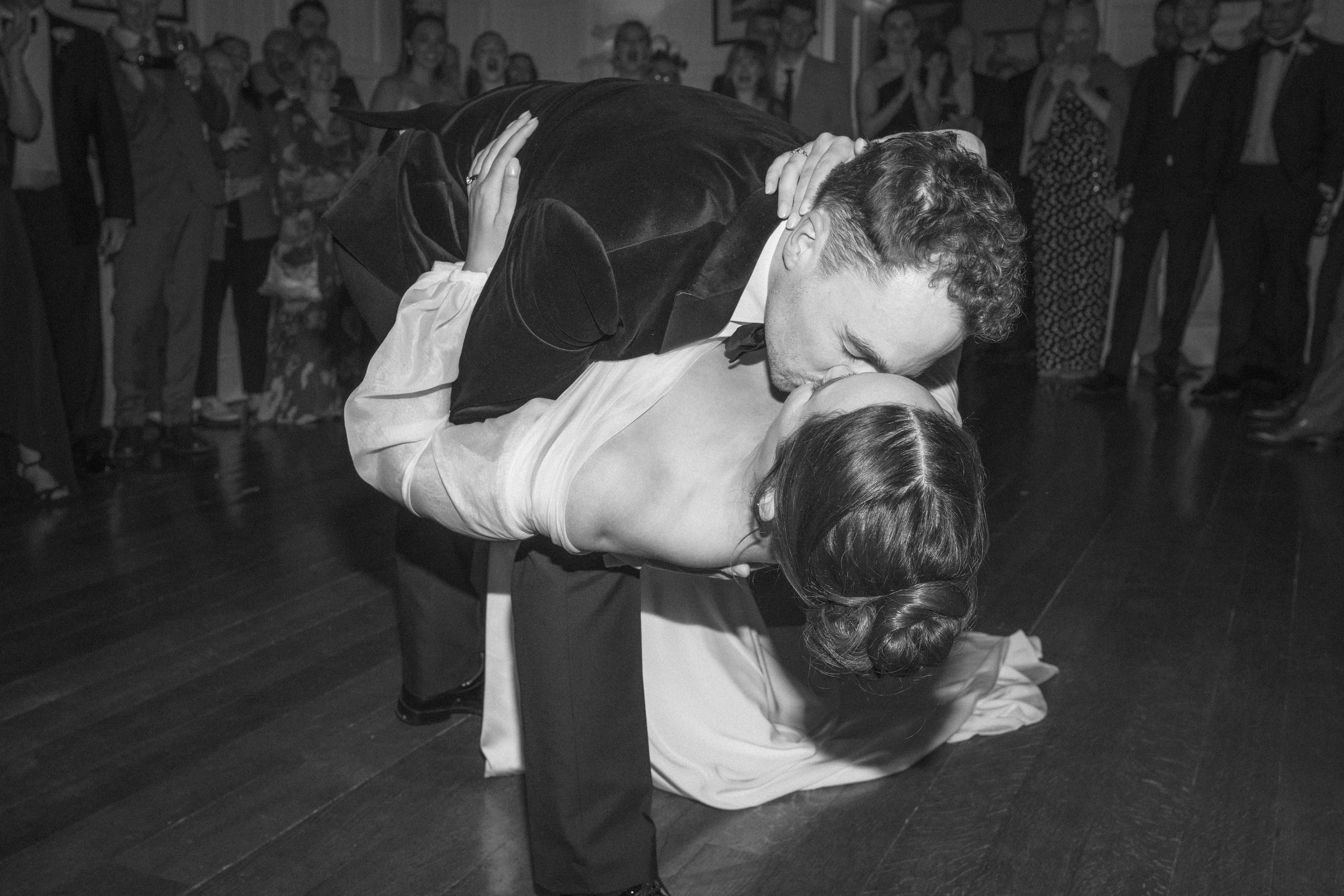 A black and white photo of a man and woman dancing closely, with the man dipping the woman as they kiss. They are dressed in formal attire, with the woman in a gown and the man in a tuxedo. A crowd watches in the background.