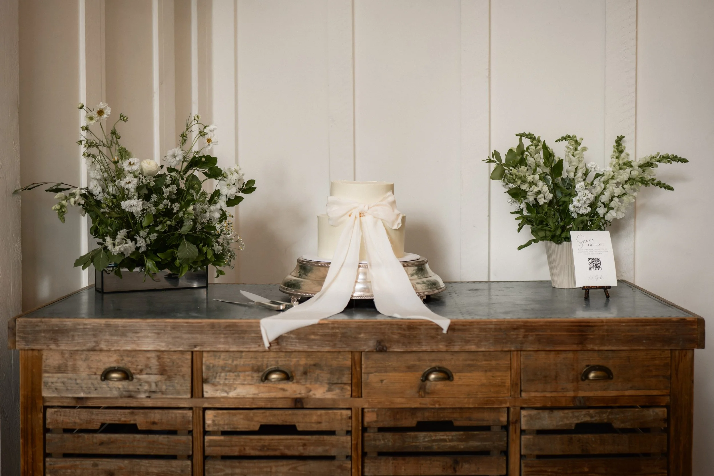 A rustic wooden sideboard with a silver cake stand, a white wedding cake with a bow, two flower arrangements in white vases, a cake knife and server, and a small sign on a stand with a QR code.