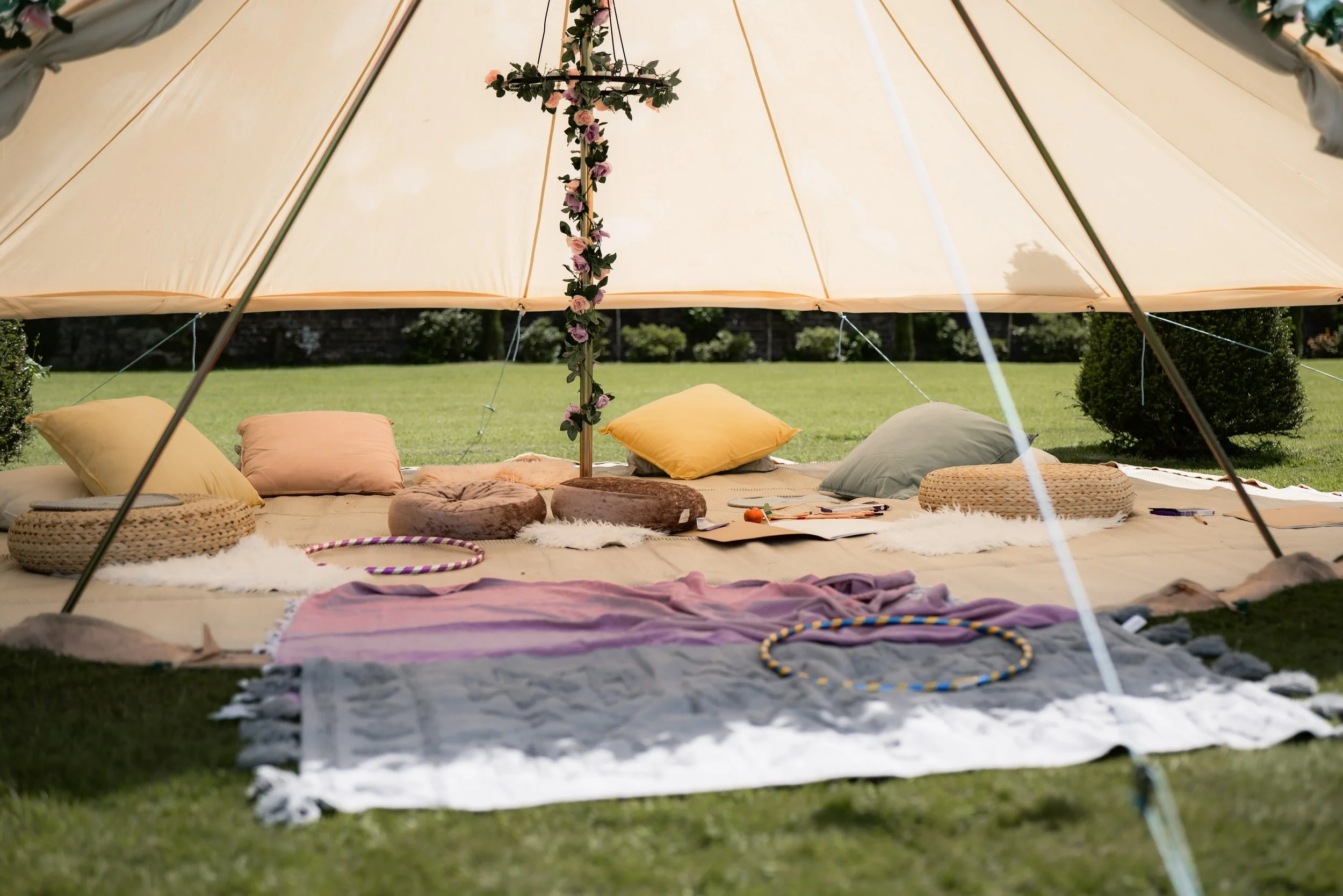 A cozy outdoor glamping tent setup with cushions, rugs, and hula hoops on a grassy field, with a tree decorated with flowers in the center.