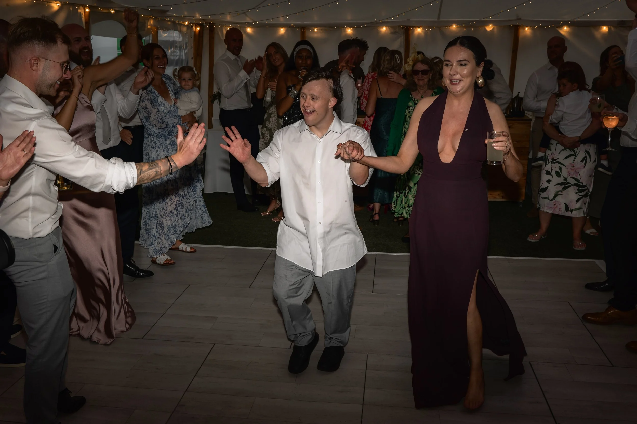 People dancing and celebrating at a party or wedding reception under string lights in a tent.