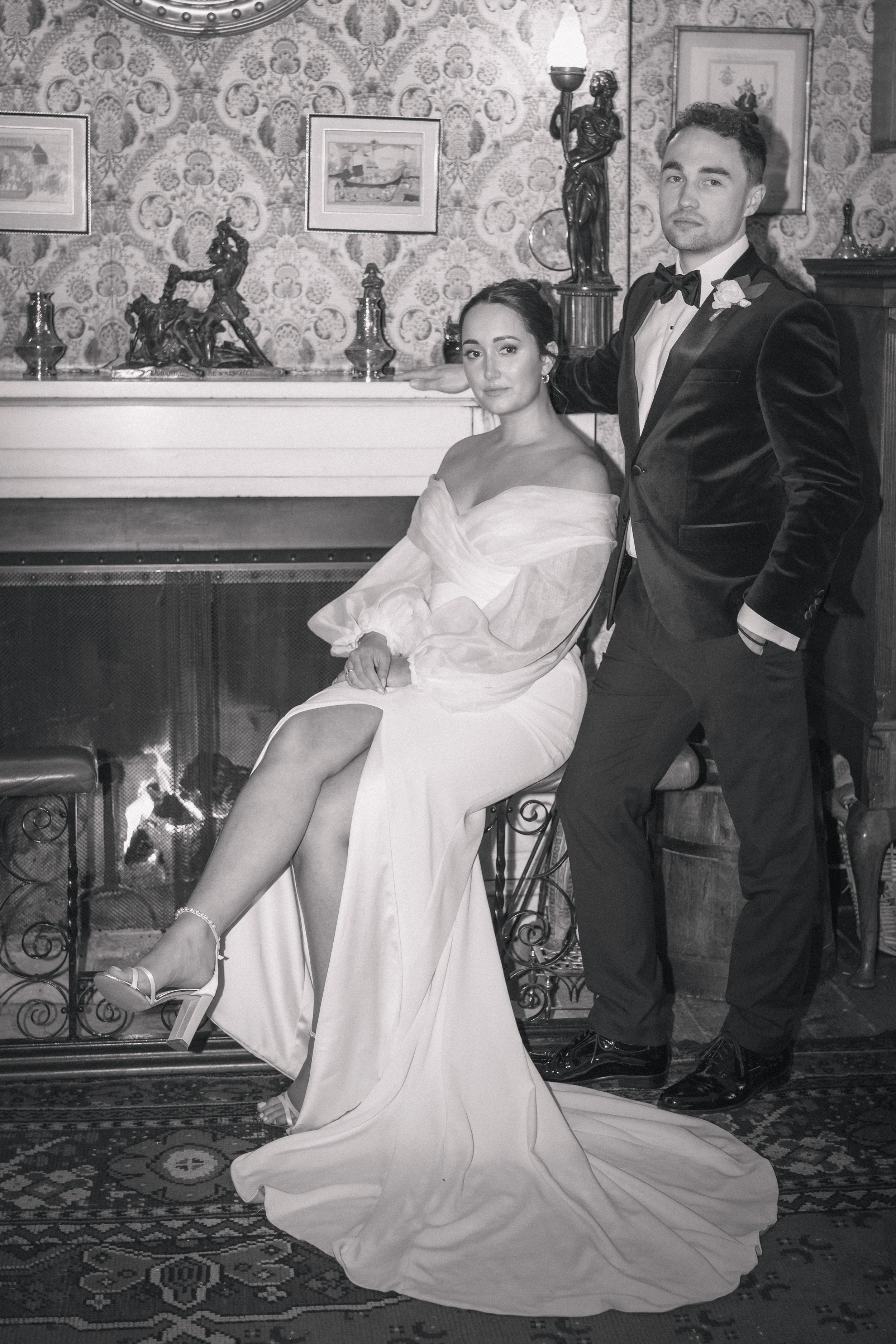 A black-and-white photo of a bride and groom posing by a fireplace in a vintage-style room. The bride is seated, wearing a white dress with her legs crossed and high heels visible. The groom stands beside her, dressed in a tuxedo with a bow tie, rest