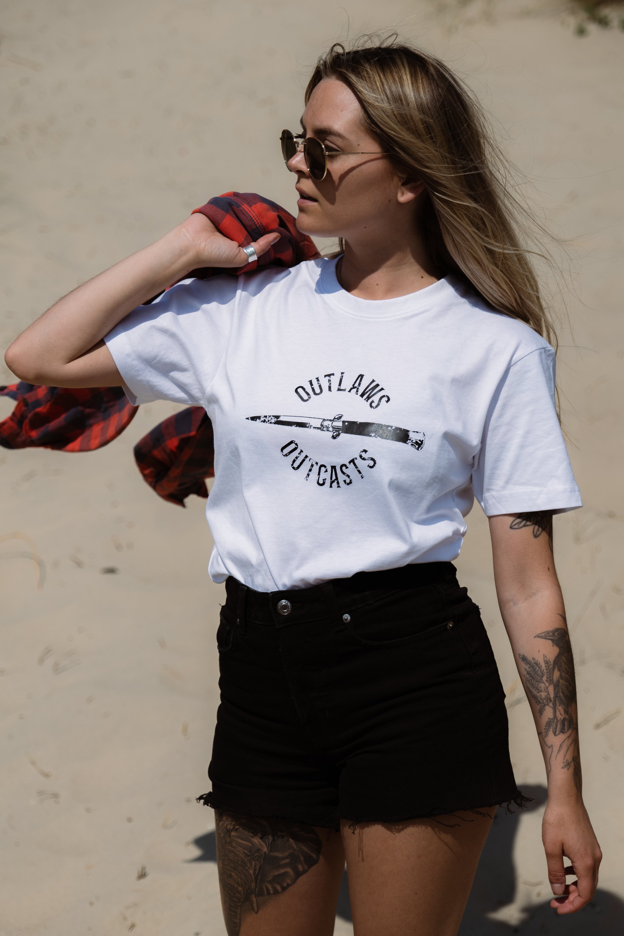 A woman with glasses and tattoos wearing a white t-shirt with a graphic and text, high-waisted black shorts, standing against a sandy background with her head turned to the side, carrying a red plaid shirt over her shoulder.