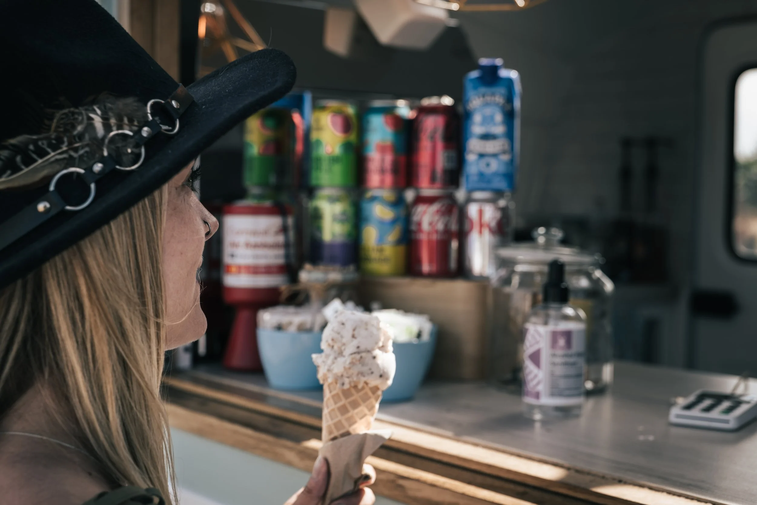 Woman with a black wide-brimmed hat eating vanilla ice cream cone inside a food truck with various canned drinks and snacks in the background.