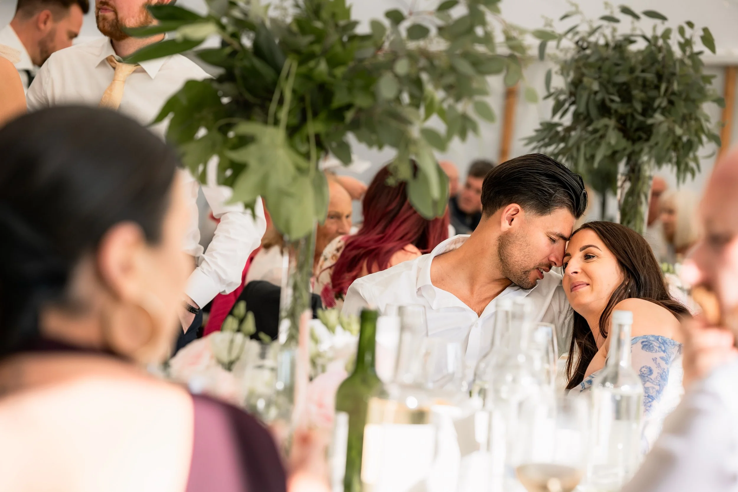 A couple shares a close moment at a wedding reception with guests and floral decorations.