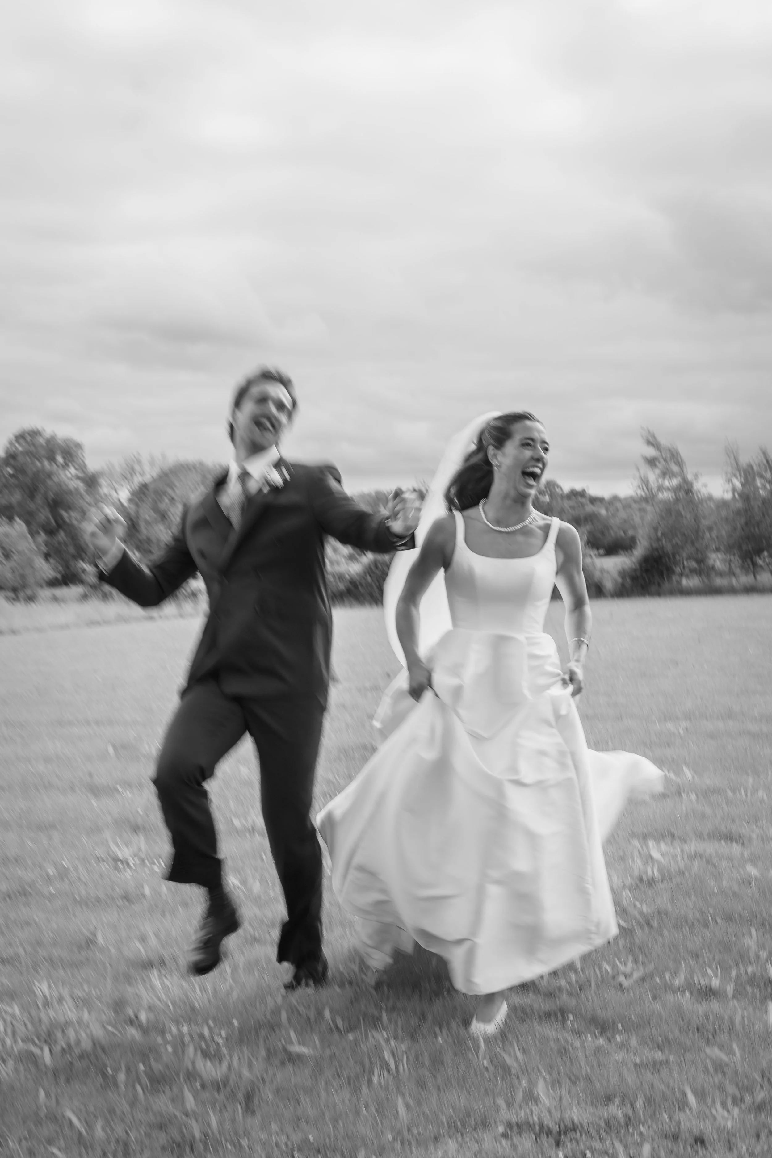 A happy couple, a bride in a wedding dress and a groom in a suit, running and laughing outdoors in a field.