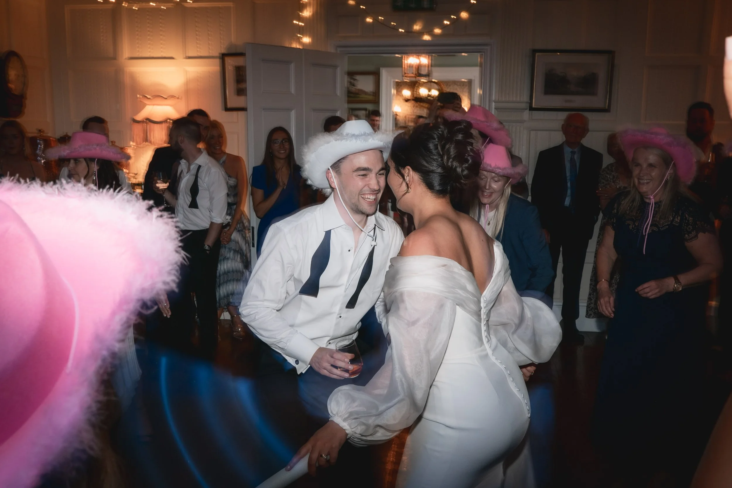 A joyful wedding reception with guests wearing pink hats, dancing, and celebrating in a decorated indoor venue.