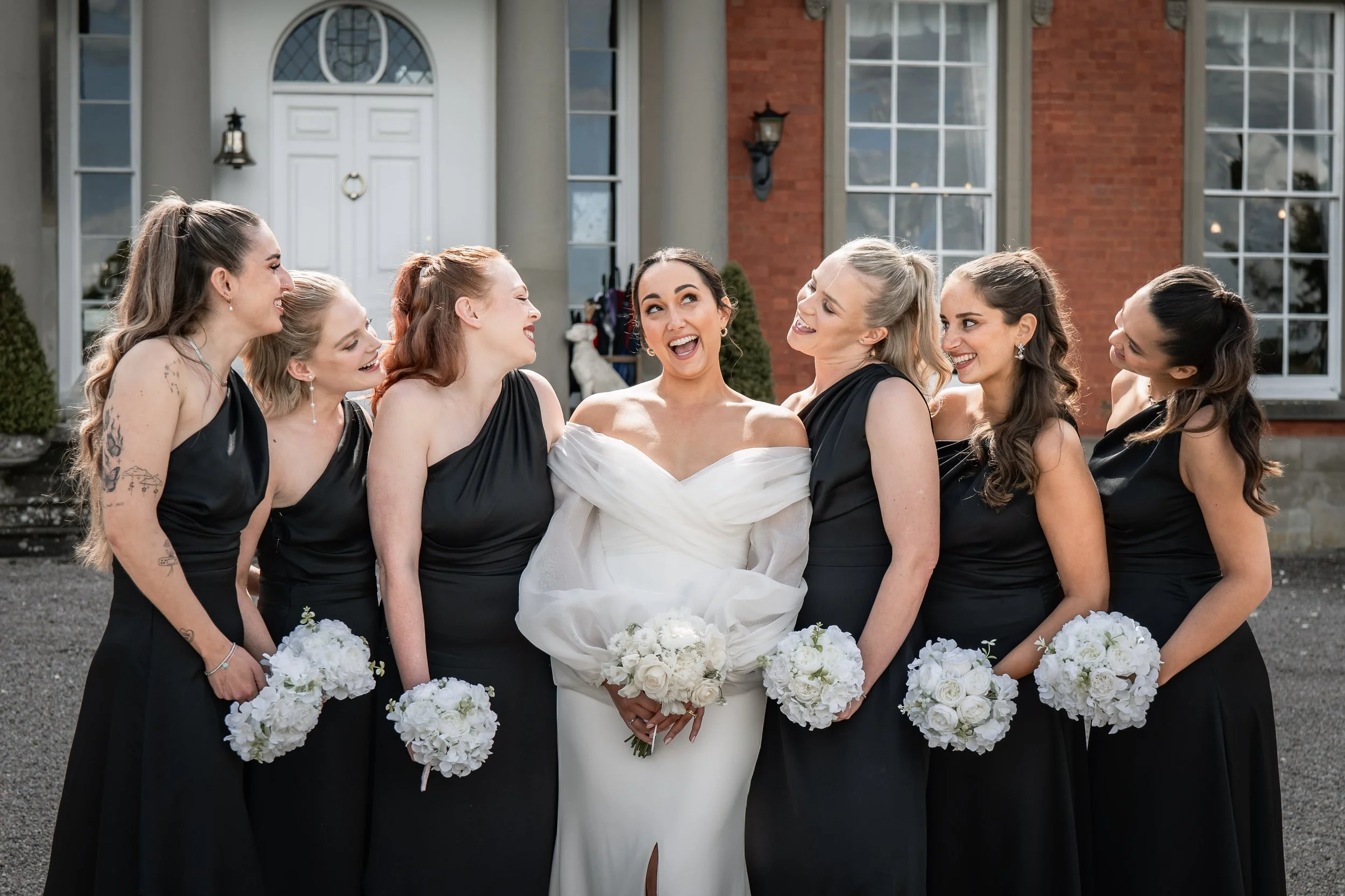Bride in a white wedding dress and six bridesmaids in black dresses standing outside a house, all holding white bouquets, smiling and laughing together.