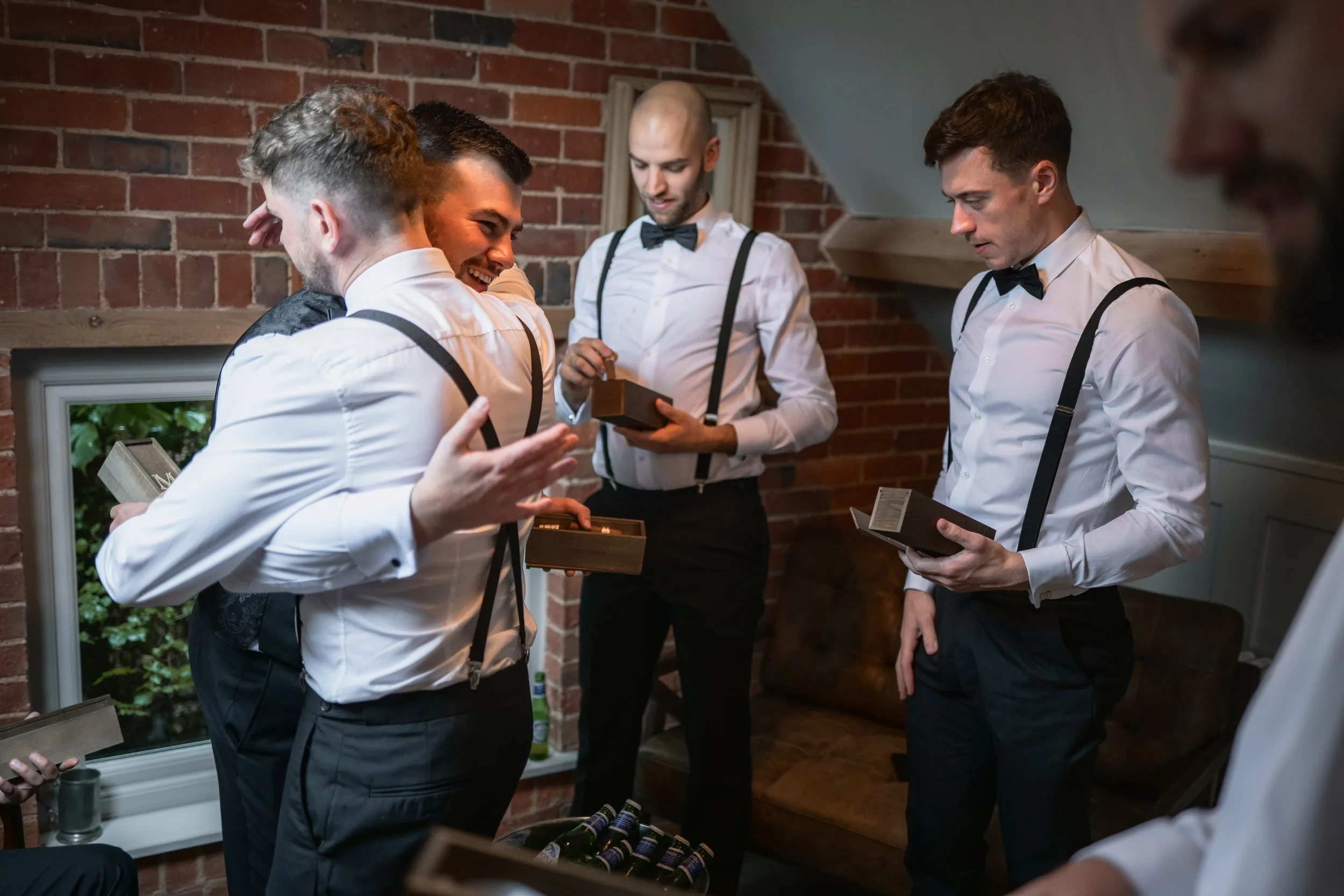 Group of men dressed in white shirts with black suspenders and bow ties gathering inside a brick-walled room, exchanging gifts or cards with joyful expressions.