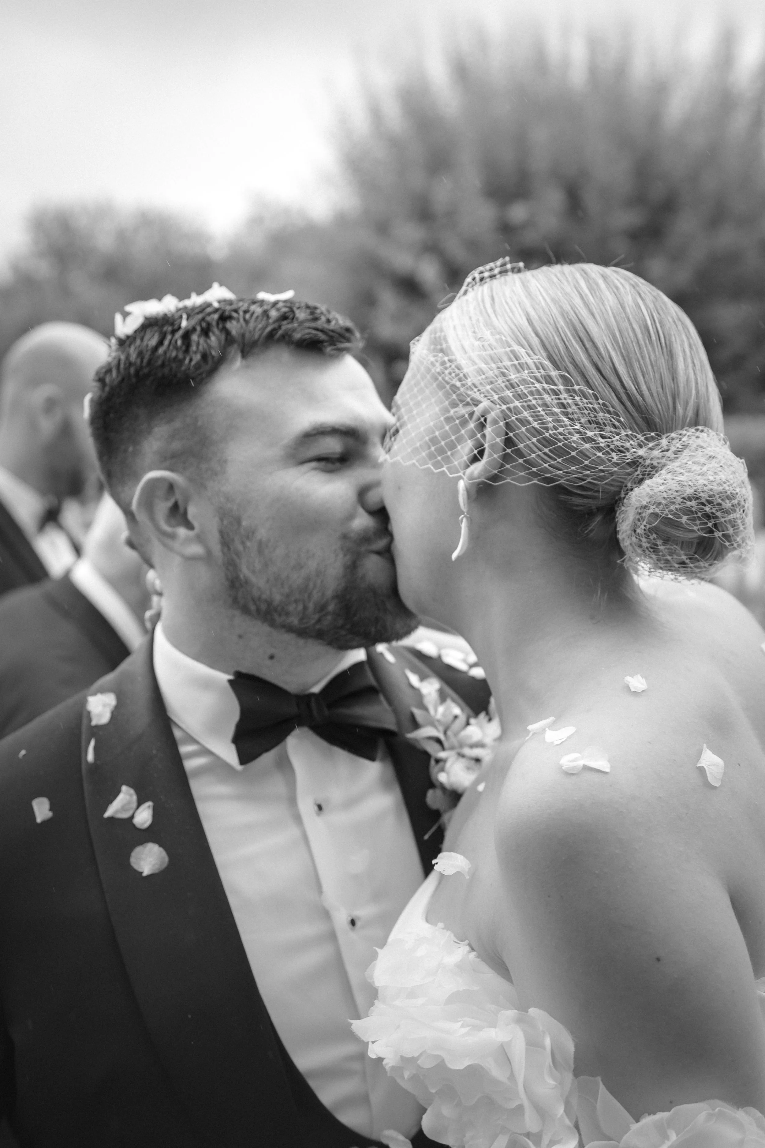 Black and white photo of a couple kissing at their wedding, with the groom in a tuxedo and bowtie, and the bride in a strapless dress with a birdcage veil.