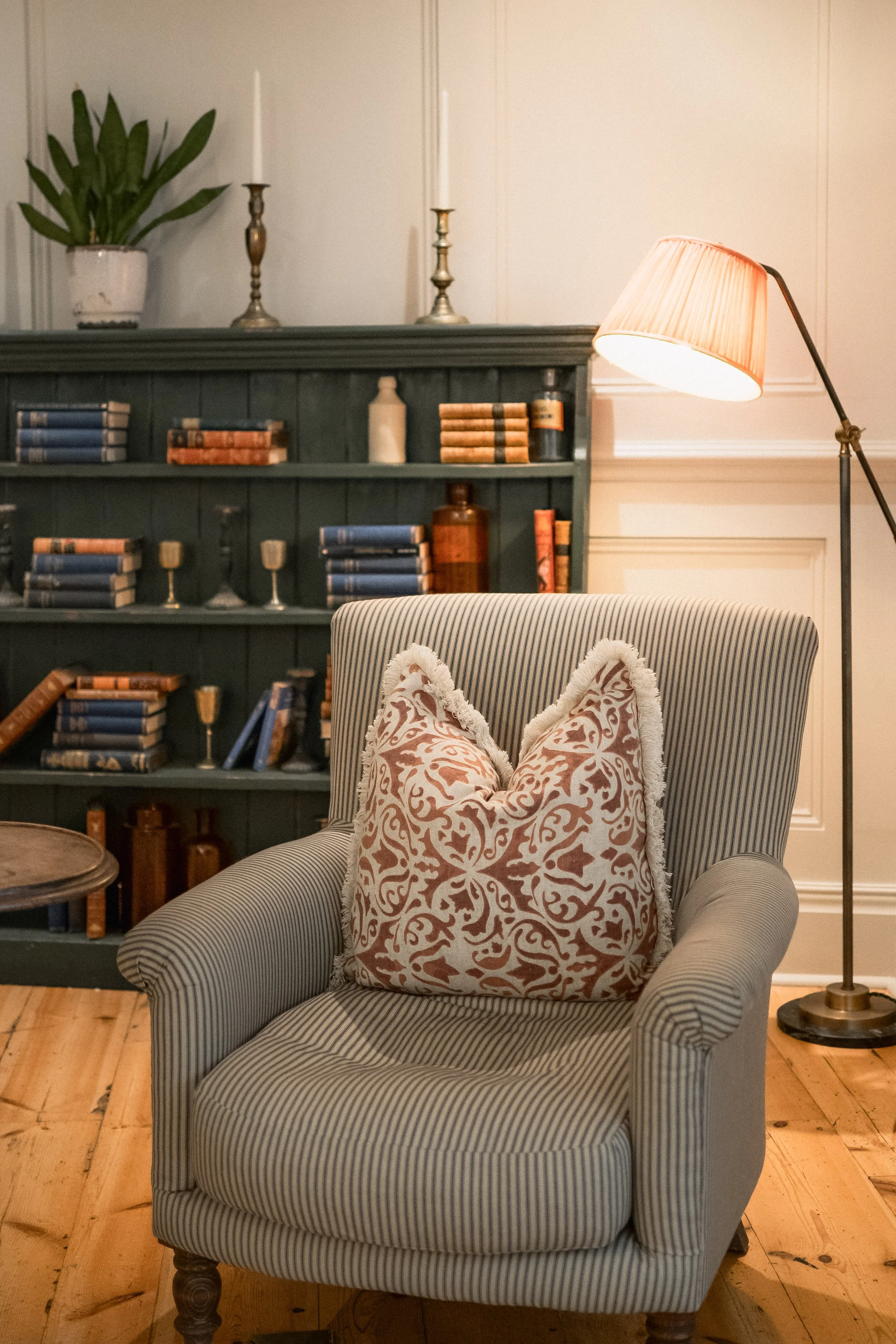 Cozy living room featuring a striped armchair with an ornate pillow, a green bookshelf filled with books and decorative items, a floor lamp with a beige shade, and a wooden floor.