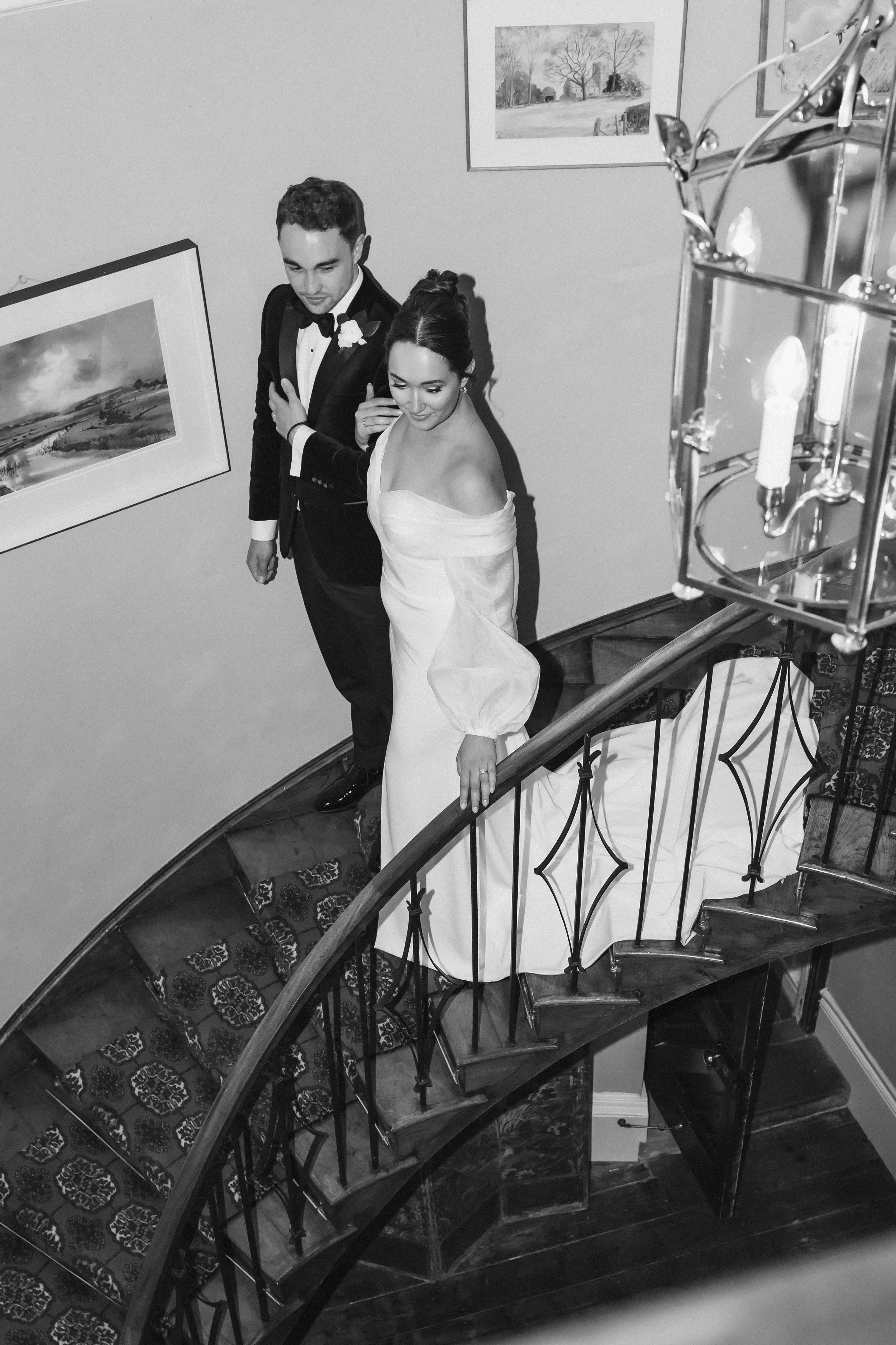 Black and white photo of a bride in a wedding dress and a groom in a tuxedo walking down a staircase.