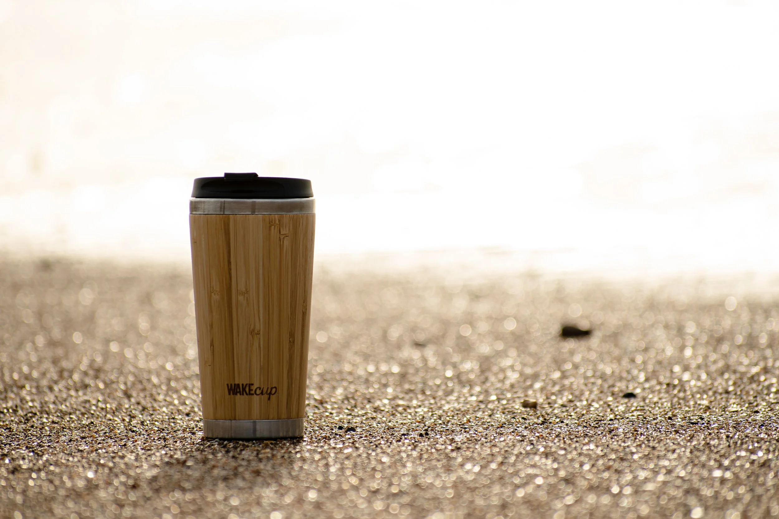 A reusable travel mug with a bamboo exterior and a black lid, standing on sandy ground outdoors.