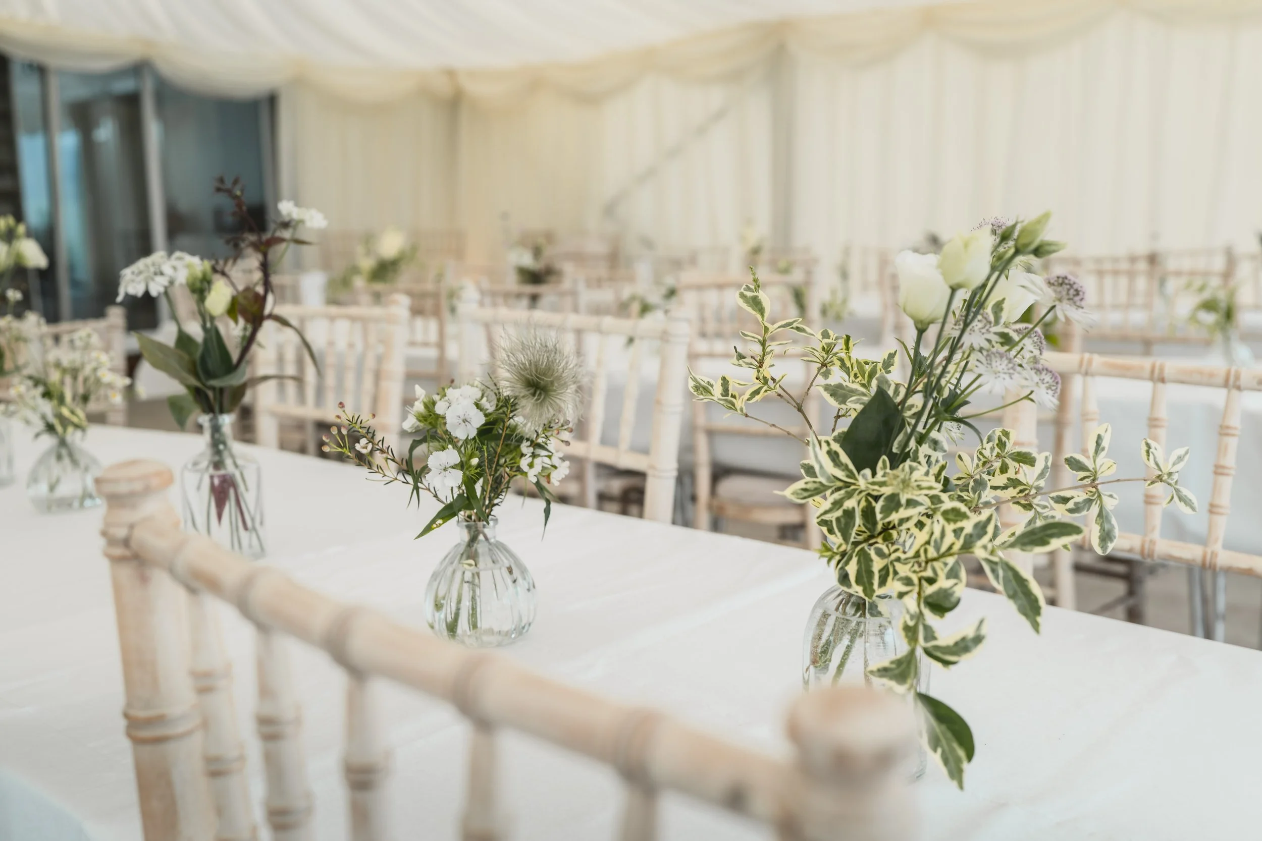 Indoor event space decorated with white and green floral arrangements in small glass vases on a white table, with wooden chairs in the background.