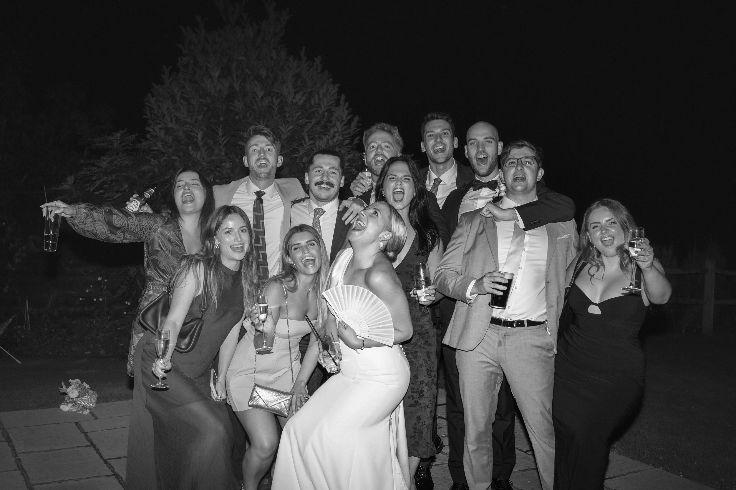 Black and white photo of a group of people celebrating, smiling, with drinks in hand, outdoors at night.