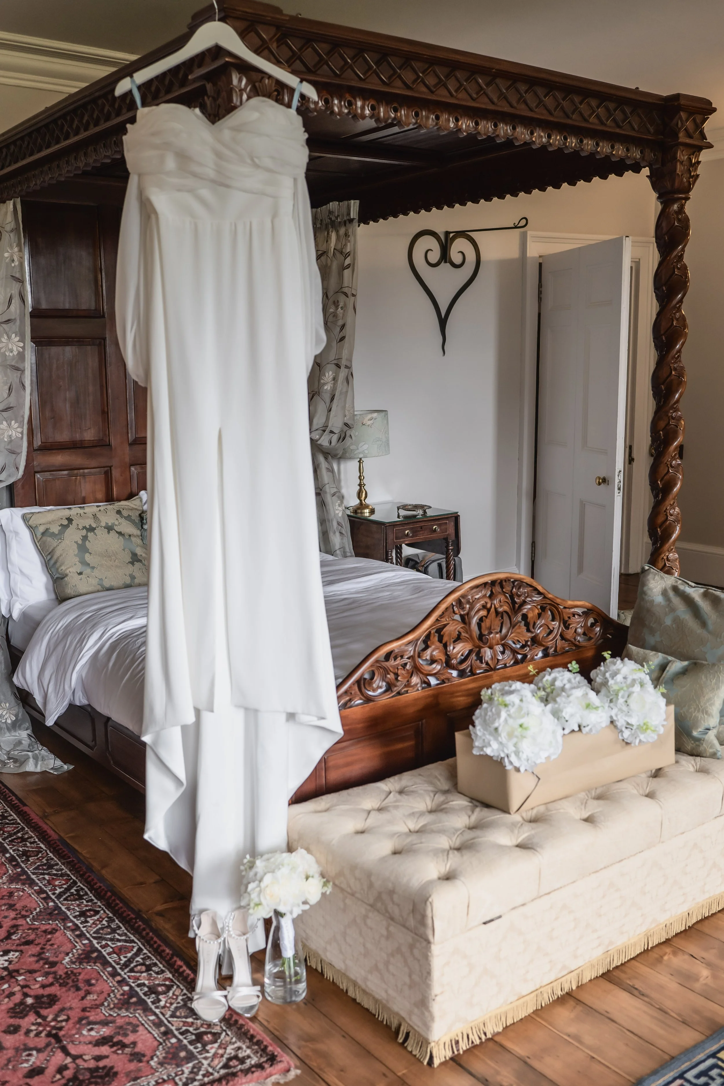 Wedding dress hanging from a canopy bed with ornate wood headboard in a bedroom decorated with floral curtains, pillows, a bedside lamp, and flowers on a bench.