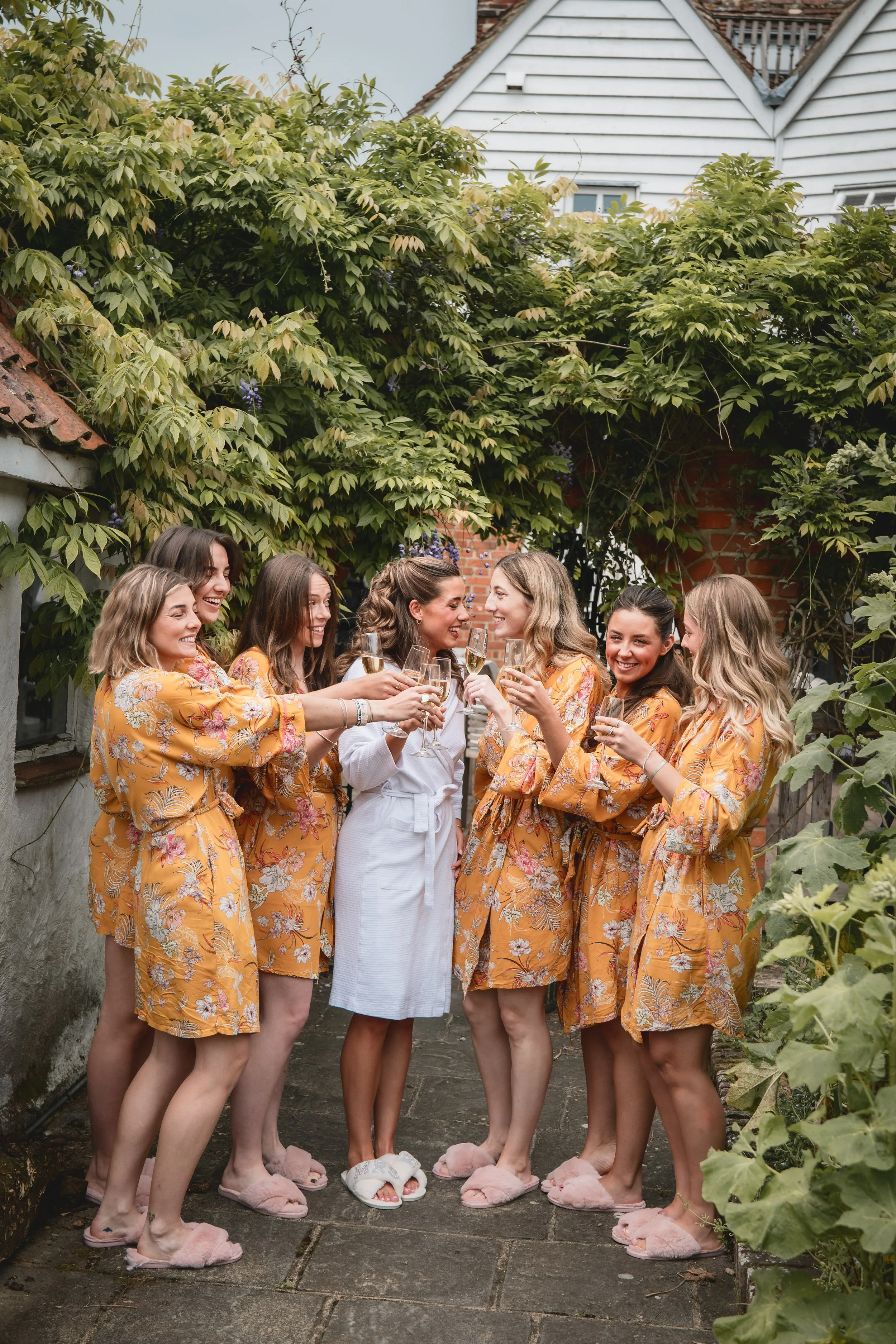 A group of seven women in matching yellow floral robes and pink slippers, holding glasses of champagne and smiling, gathered outdoors on a stone patio with lush green bushes and a brick wall in the background.