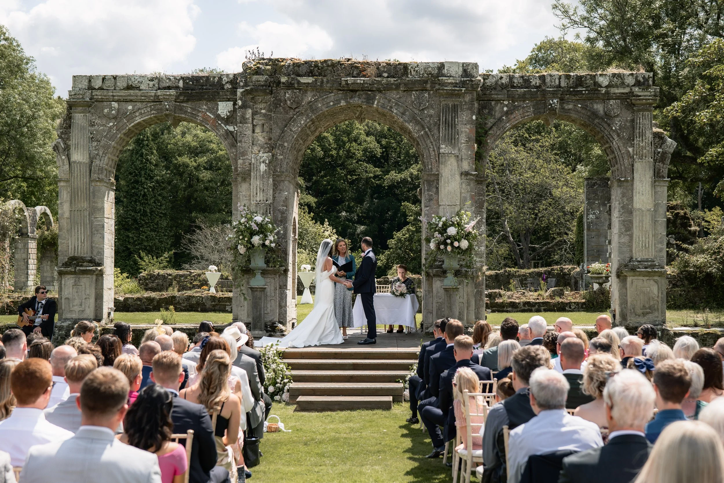 Outdoor wedding ceremony featuring bride and groom standing under ancient stone archway, exchanging vows, with officiant present. Guests seated on green lawn, surrounded by trees, with floral arrangements and musicians.