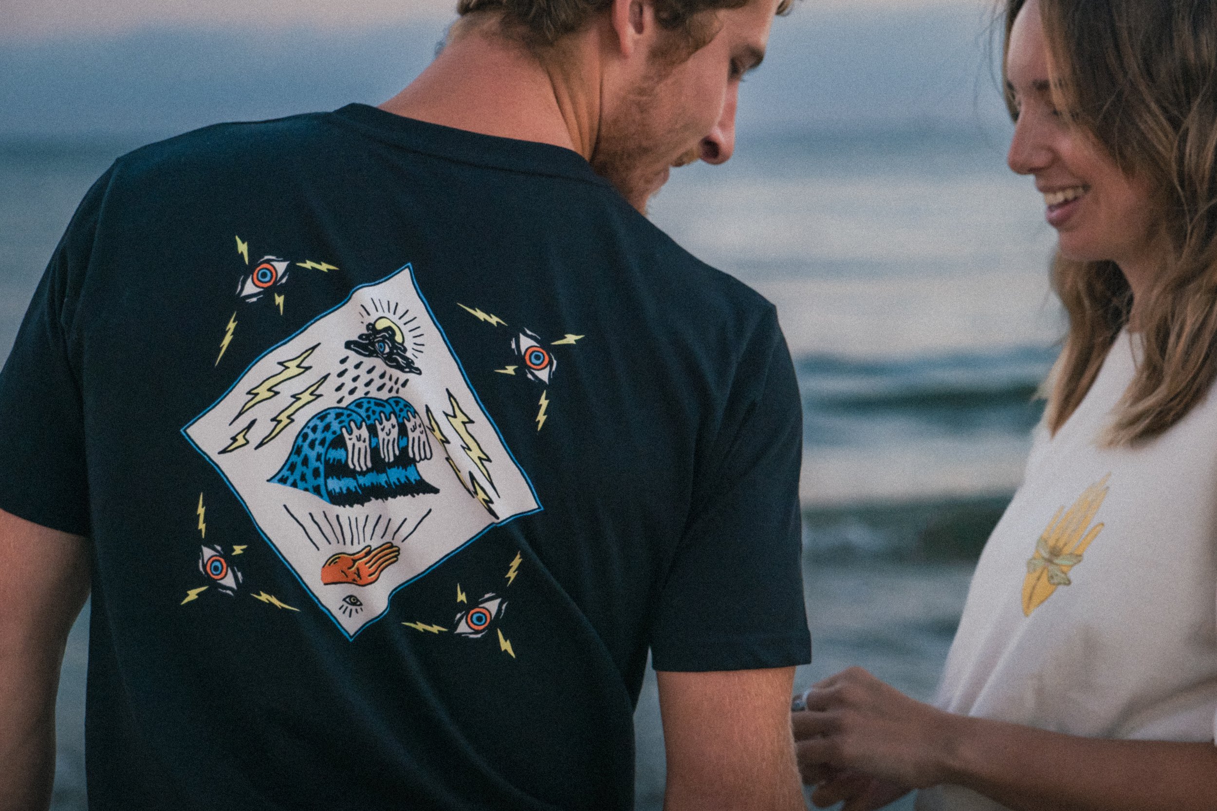 A man and woman standing close together at the beach, with the ocean in the background, smiling at each other. The man is wearing a black T-shirt with a colorful graphic on the back, and the woman is wearing a light-colored shirt with a small embroid